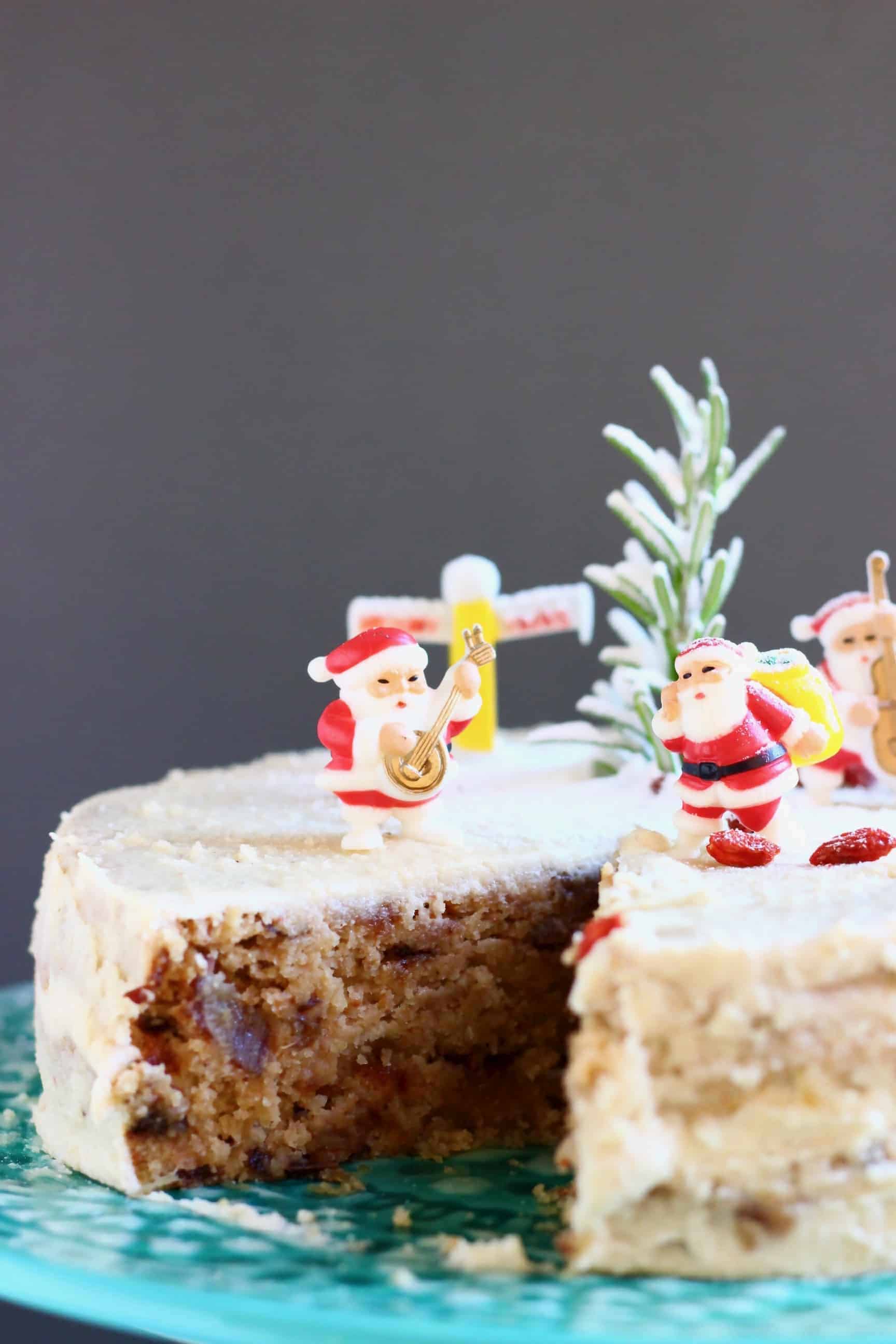 A fruit cake covered in white buttercream with a slice taken out of it topped with plastic santas against a grey background