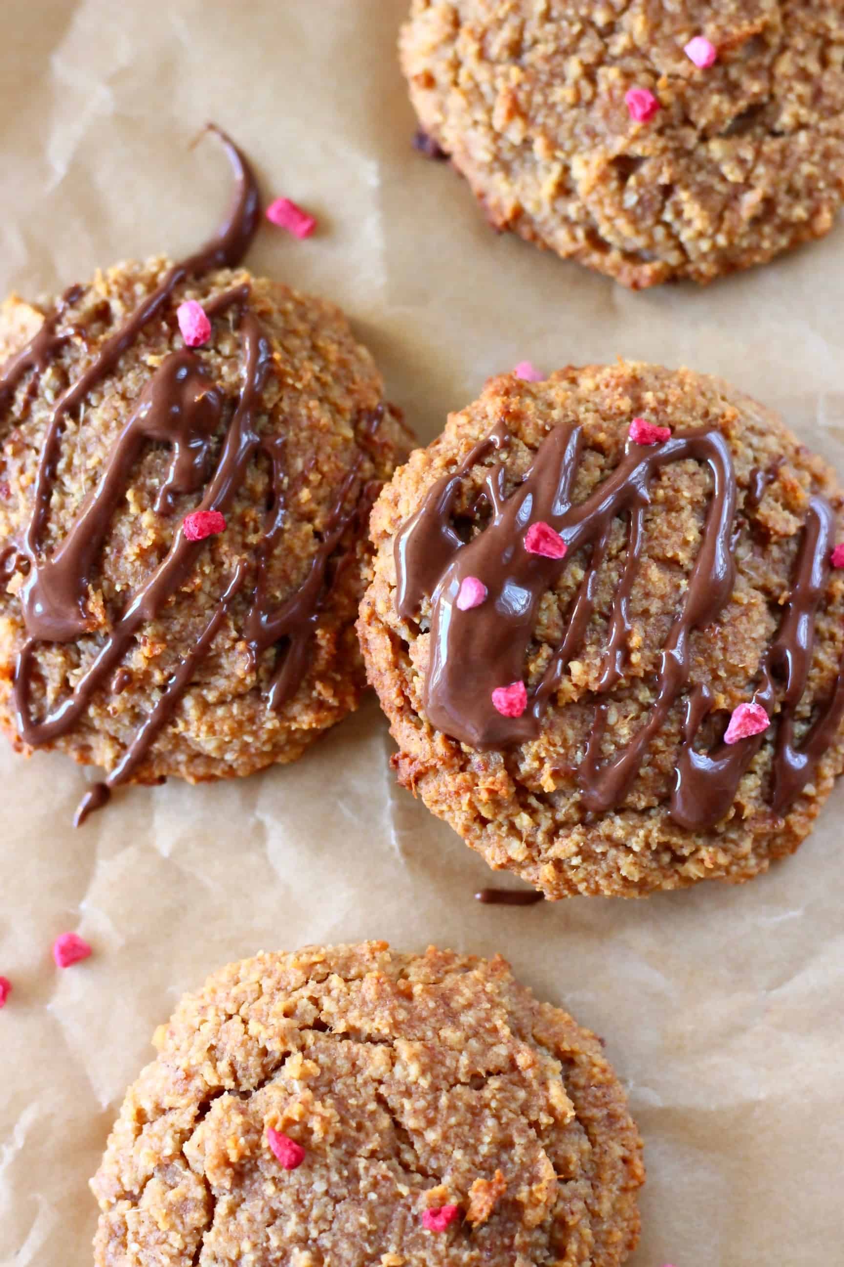 Four ginger cookies on a sheet of brown baking paper with two drizzled with melted dark chocolate and sprinkled with freeze-dried raspberries