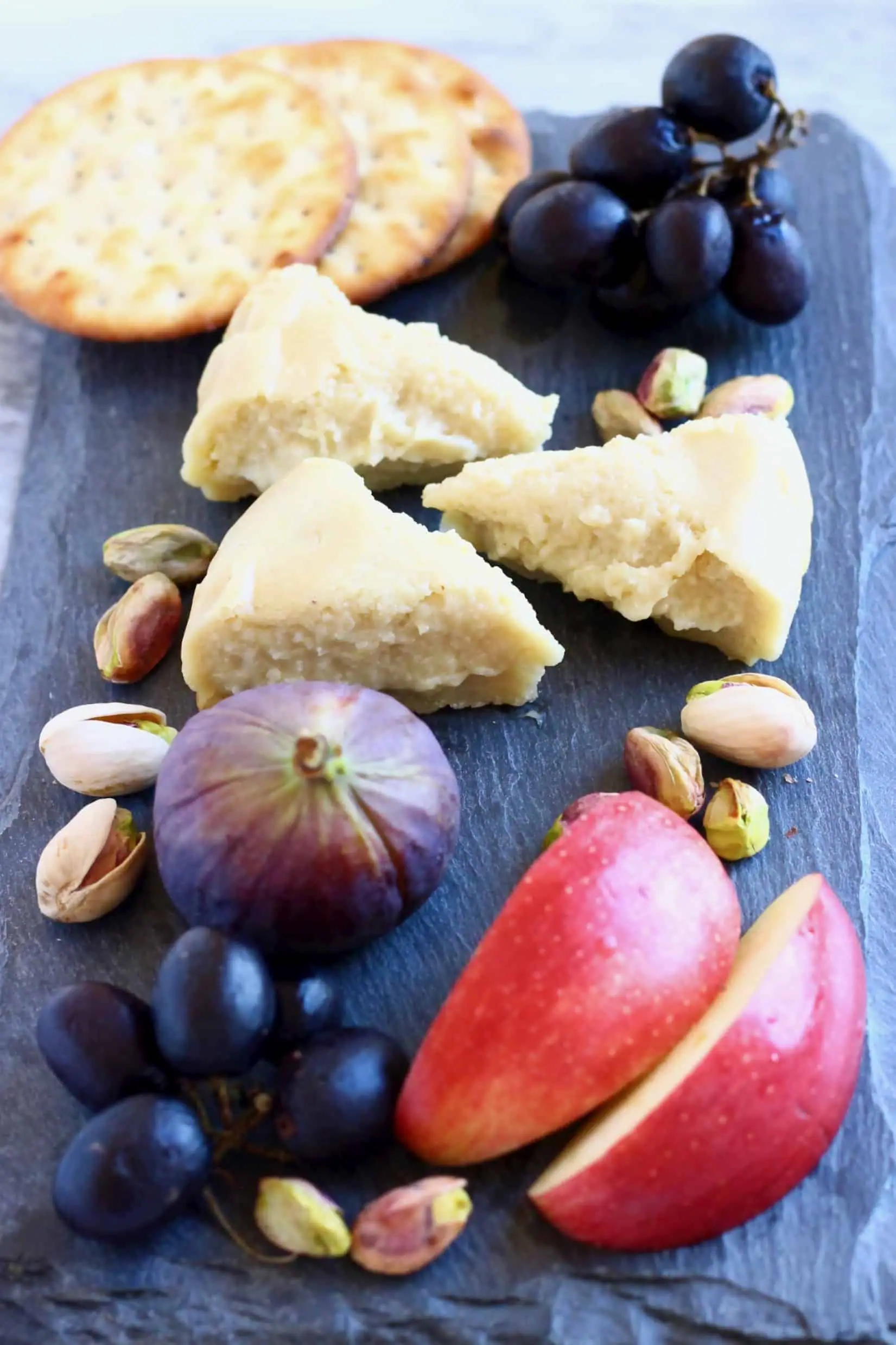 Three triangular slices of brie on a black slab with sliced apples, grapes, figs, pistachios and crackers