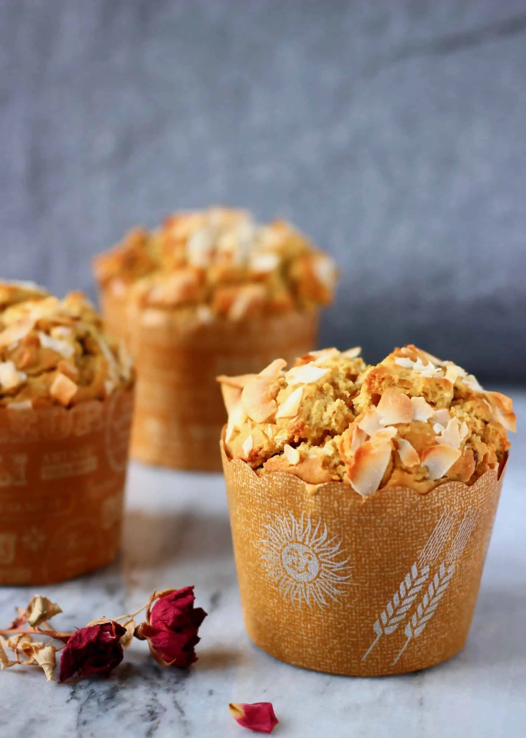 Three carrot muffins topped with coconut flakes on a marble slab decorated with dried roses against a grey fabric background