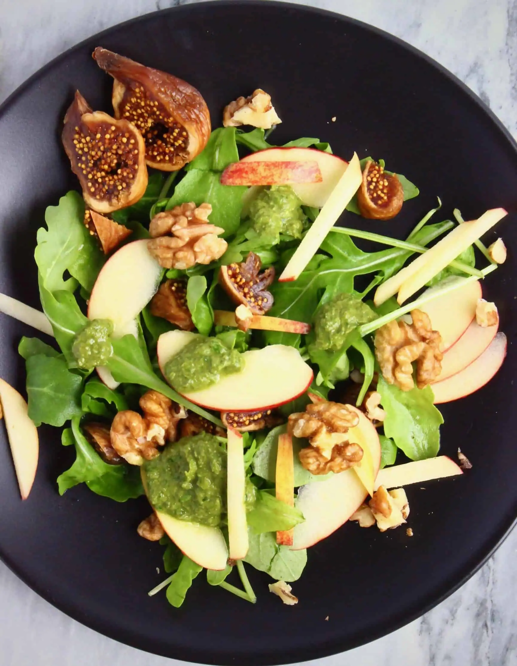 Apple slices, chopped dried figs, rocket and walnuts covered in a green pesto dressing on a black plate against a marble background