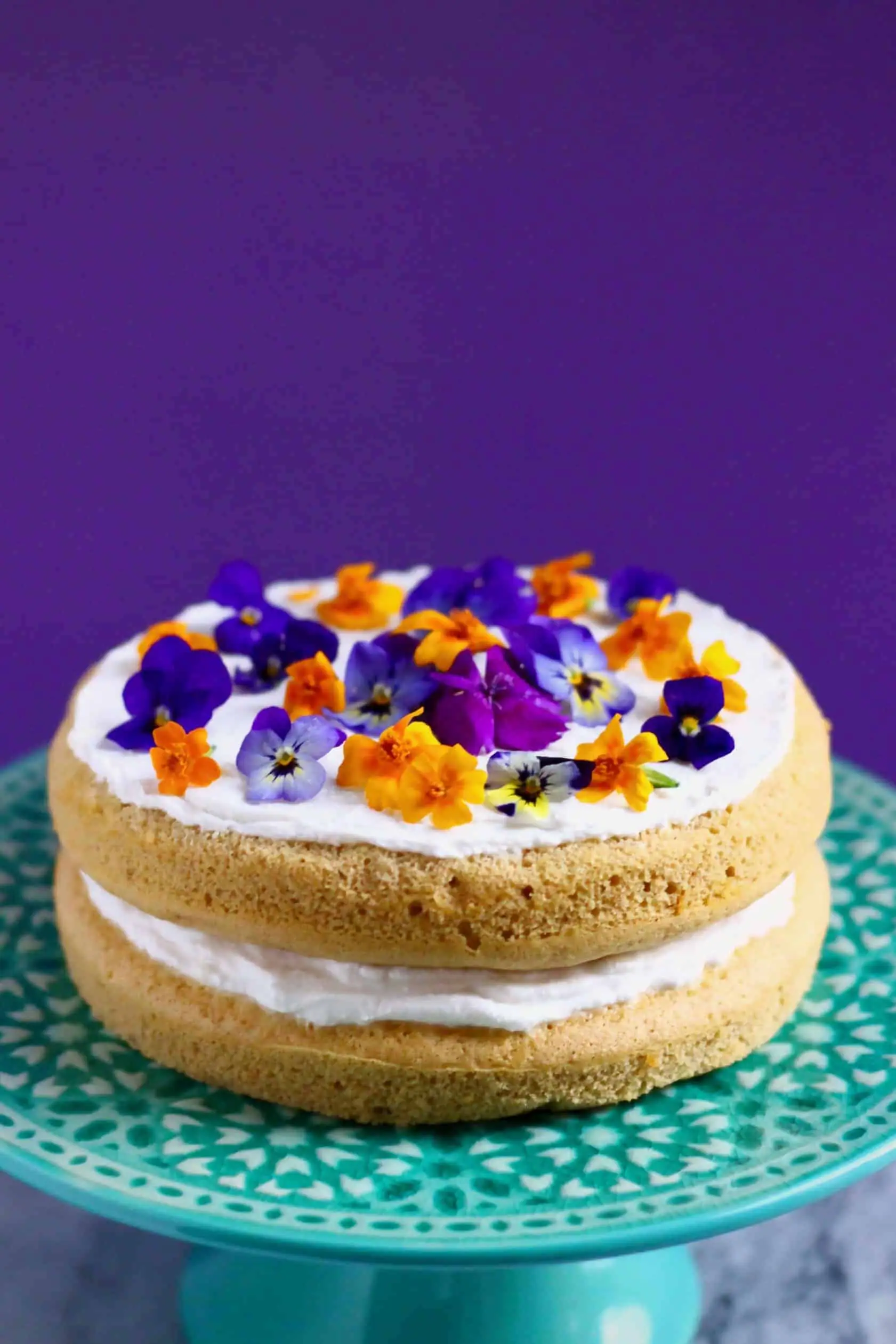 A pumpkin sponge sandwiched with white frosting topped with purple and orange flowers on a cake stand 