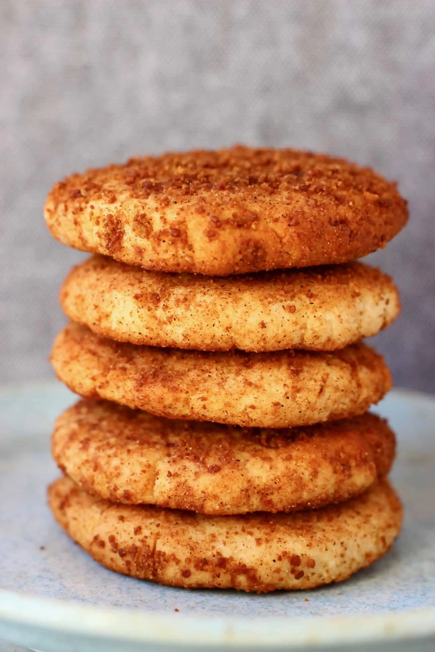 Five cookies coated in brown cinnamon sugar stacked on top of each other on a light blue plate against a grey background