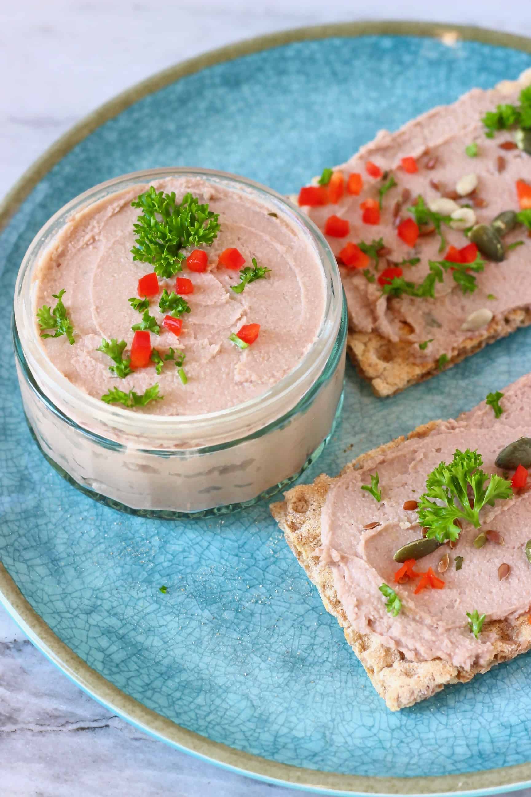 A glass pot filled with beige pâté topped with herbs and chopped red pepper on a blue plate with two rectangular crackers spread with pâté and decorated with pumpkin seeds, chopped red chilli and green herbs