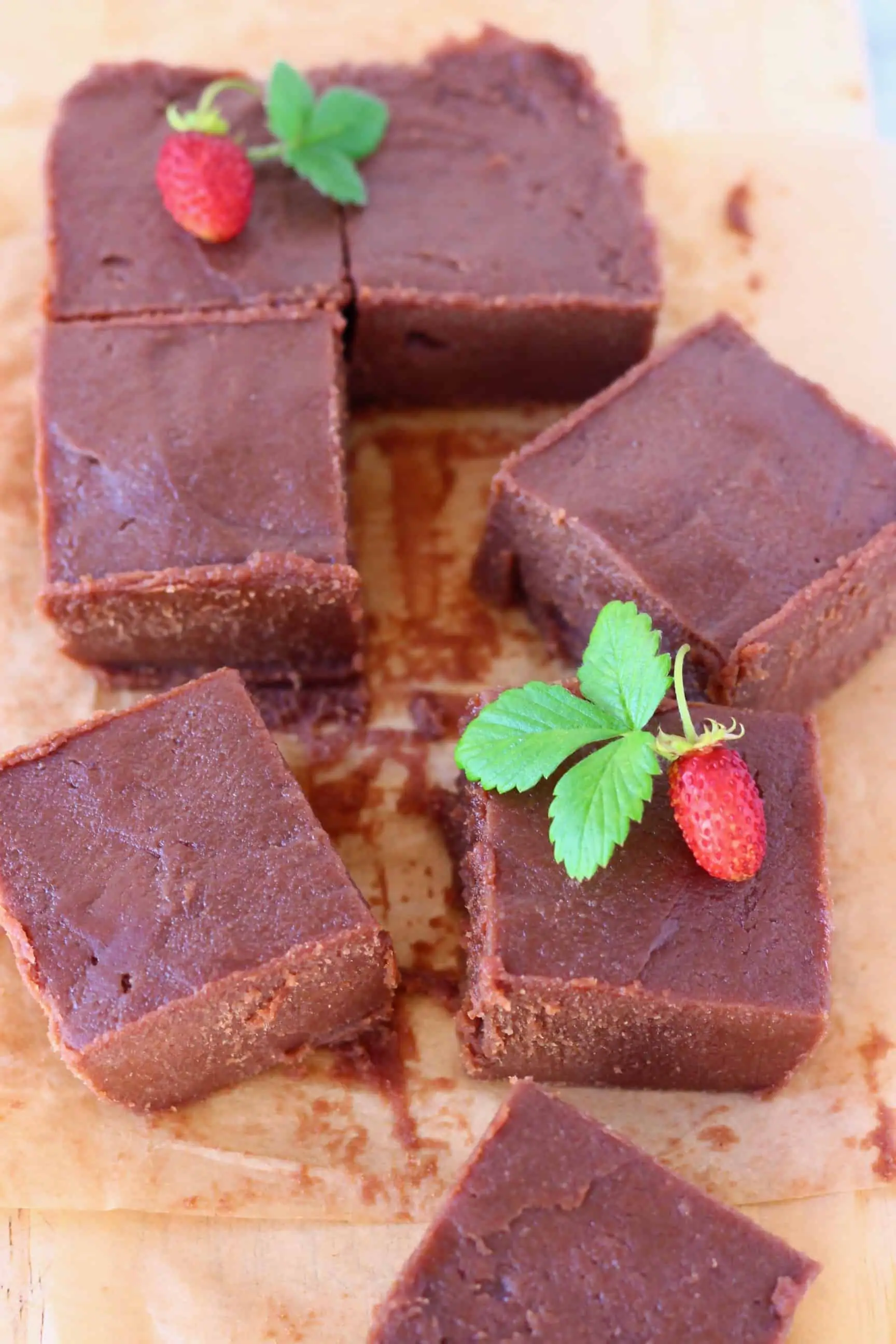 Seven square pieces of chocolate fudge on a sheet of brown baking paper decorated with mini strawberries with green leaves