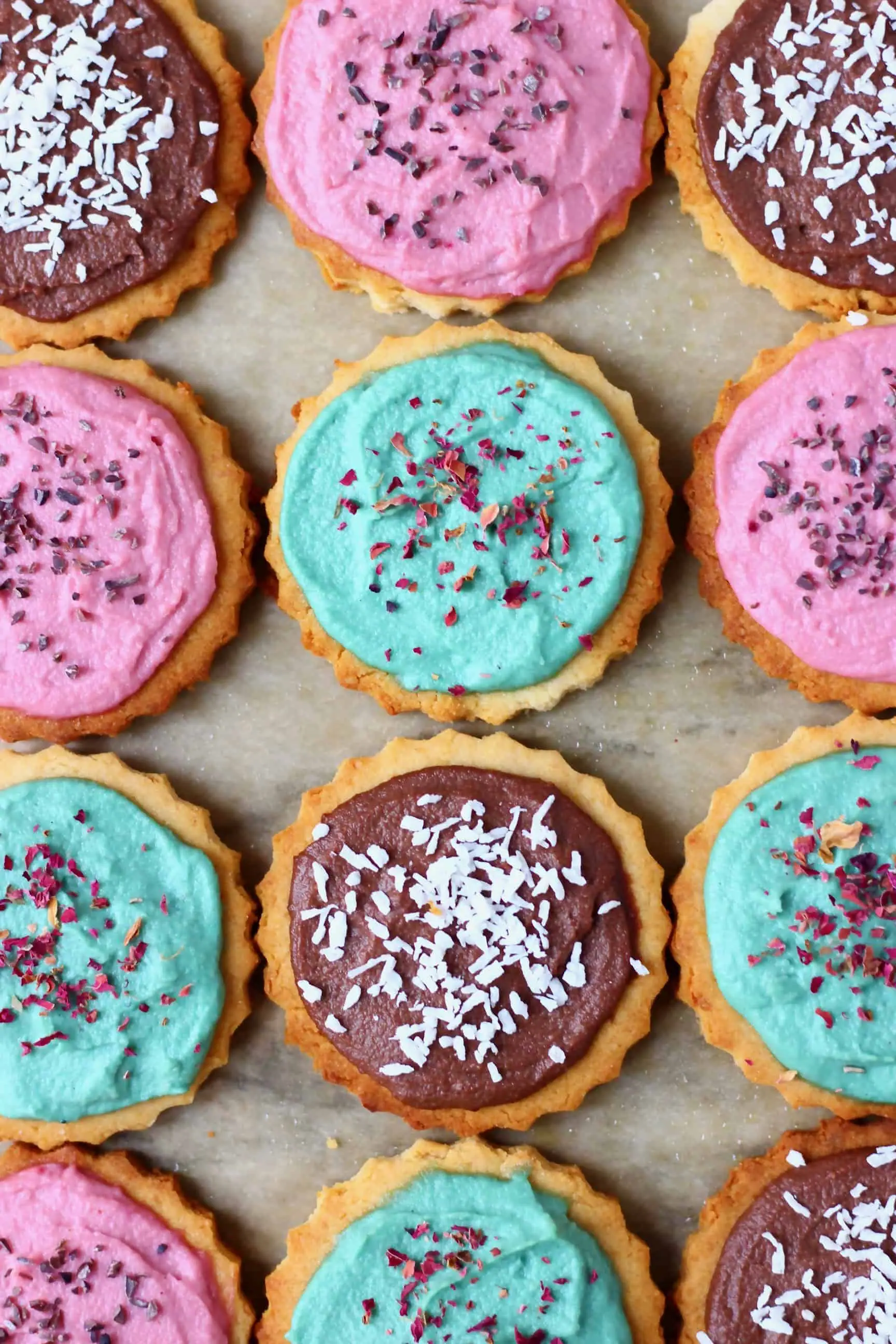 Twelve sugar cookies topped with different coloured frosting and sprinkles on a sheet of brown baking paper