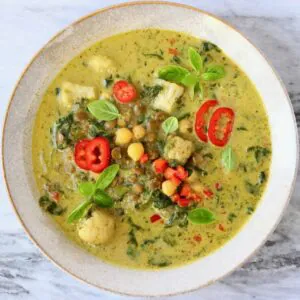 Photo of green curry with cauliflower, spinach, chickpeas and green lentils topped with basil and red chilli in a beige bowl against a marble background