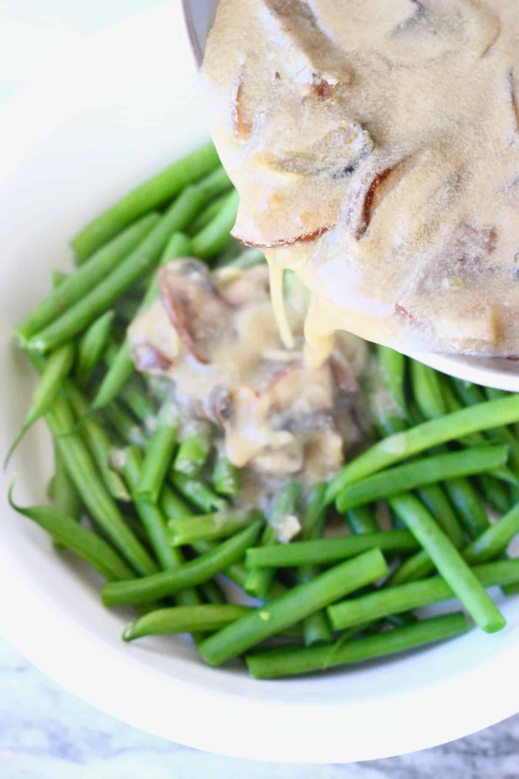Green beans in a pie dish with a pan pouring over sliced mushrooms in a creamy brown sauce