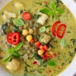 Photo of green curry with cauliflower, spinach, chickpeas and green lentils topped with basil and red chilli in a beige bowl against a marble background