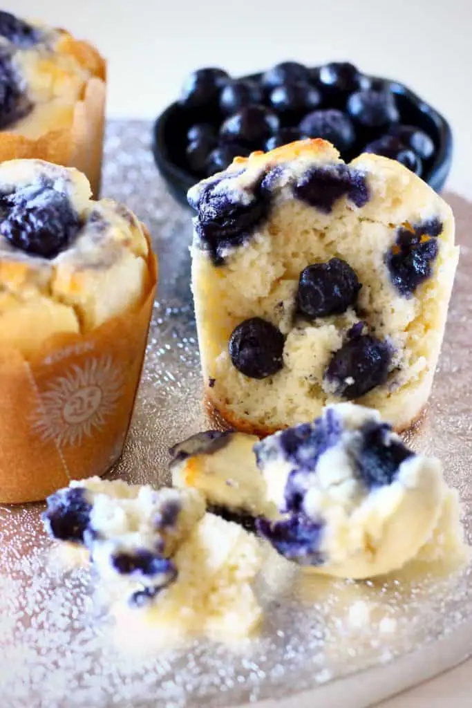 Photo of three blueberry muffins, one with a bite taken out of it, with a bowl of fresh blueberries in the background