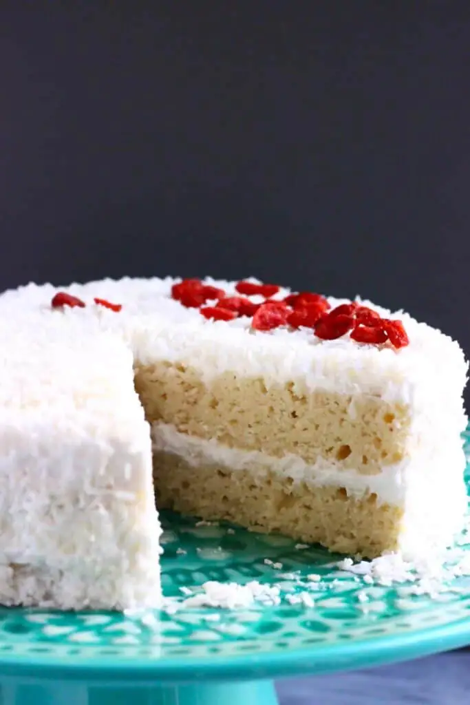Photo of a white sponge cake covered in creamy frosting and coconut sprinkled with red goji berries against a dark grey background