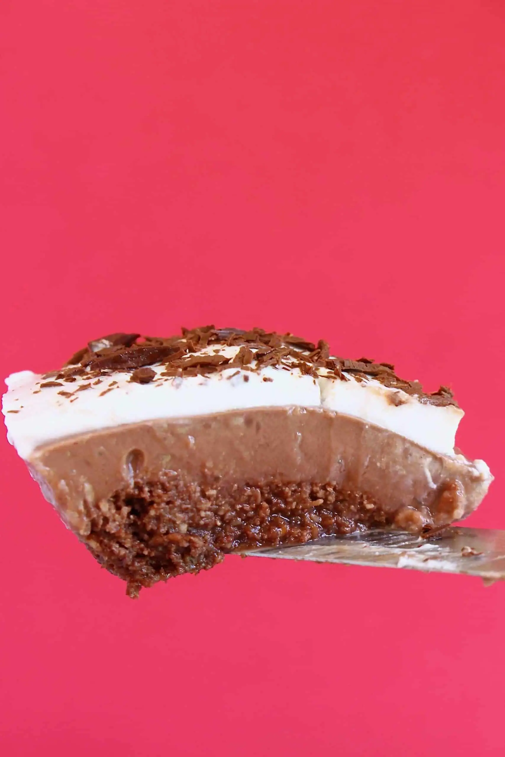 Photo of a slice of chocolate pie topped with whipped cream and chocolate shavings being held up against a bright pink background
