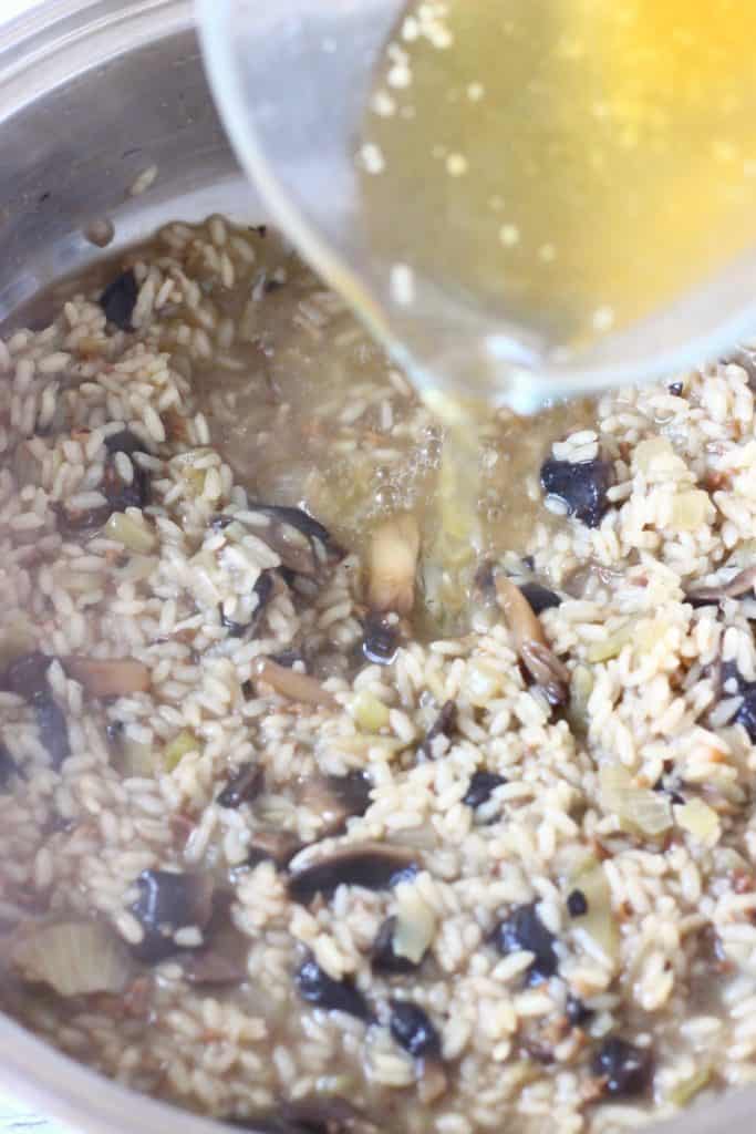 Sliced mushrooms and rice in a pan with a jug pouring broth into it