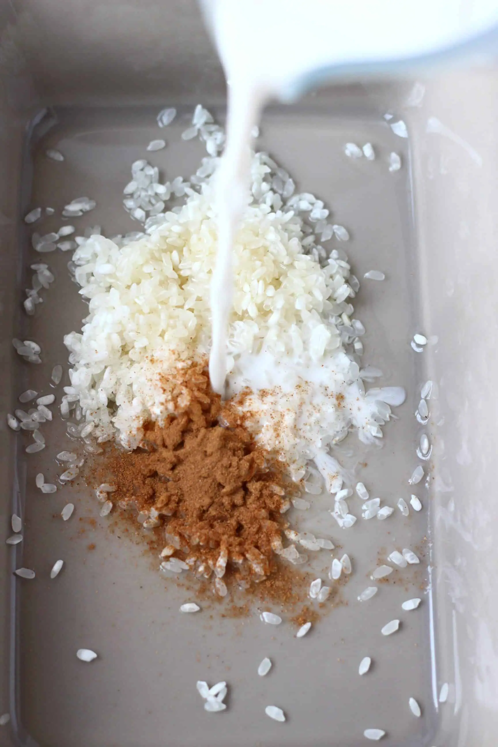 Ground cinnamon and uncooked rice in a grey baking tray with milk being poured into it in a jug