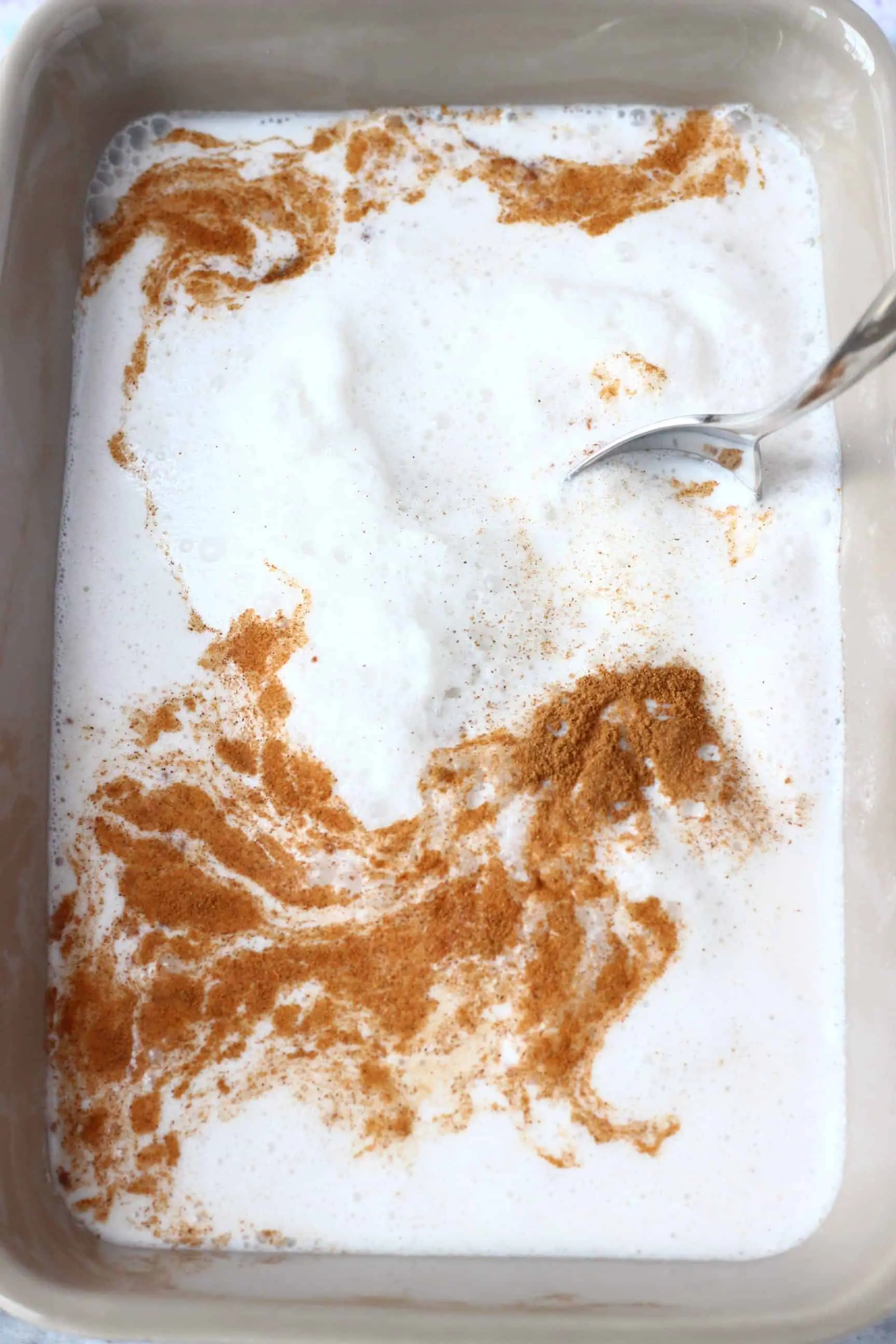 A grey baking tray with rice, cinnamon and milk with a silver spoon mixing it around