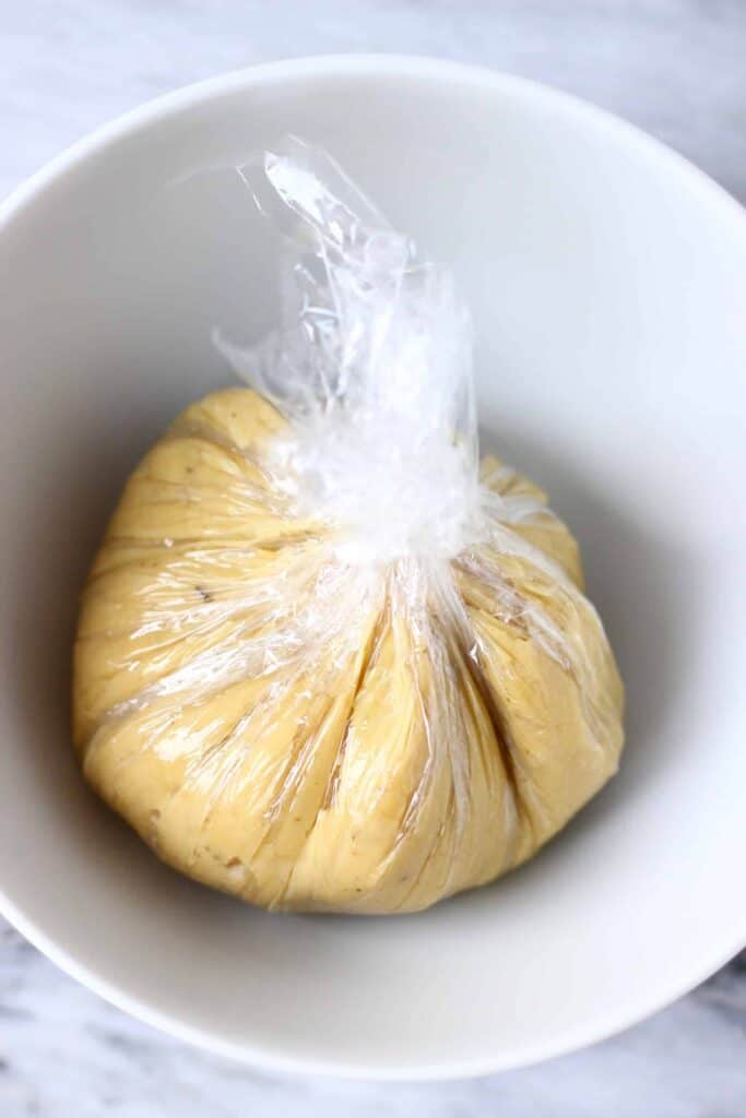 A ball of cashew cheese wrapped in cling film in a white bowl against a marble background