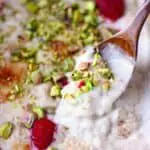 Vegan rice pudding in a grey rectangular baking dish topped with chopped pistachios and fresh raspberries with a wooden spoon lifting up a mouthful of it