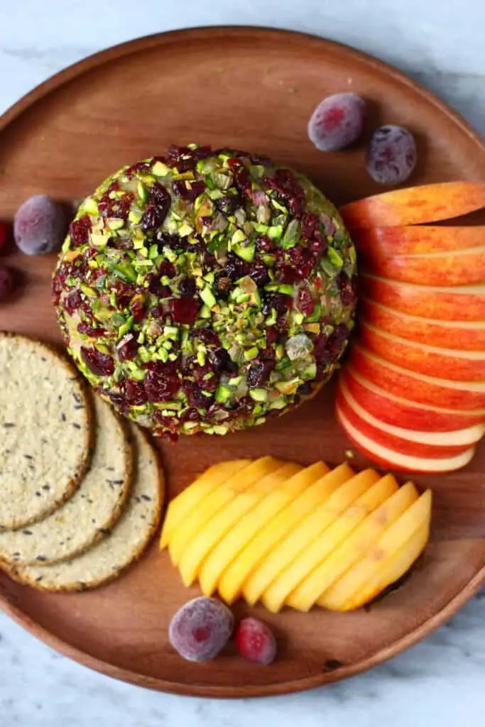 Photo of a ball of cashew cheese covered with chopped pistachios and dried cranberries on a wooden plate with sliced red apple, sliced persimmon, oatcakes and fresh cranberries