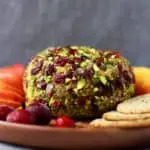 Photo of a ball of cashew cheese covered with chopped pistachios and dried cranberries on a wooden plate with sliced apples, oatcakes and fresh cranberries against a grey background