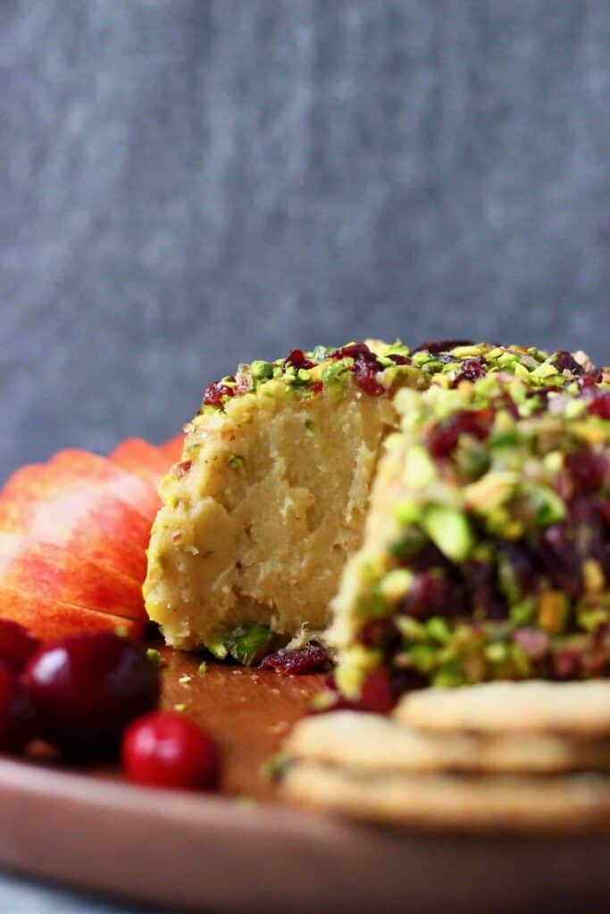 A sliced ball of cashew cheese covered in chopped pistachios and dried cranberries on a wooden plate against a grey background
