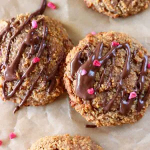 Two golden brown cookies on a sheet of brown baking paper drizzled with melted dark chocolate and sprinkled with freeze-dried raspberries