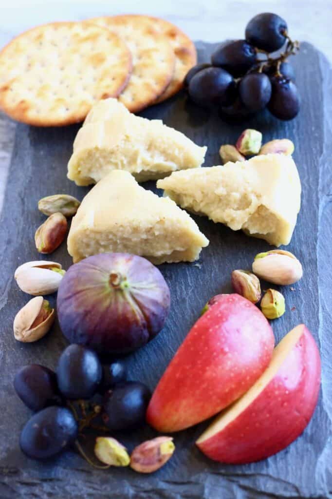 Photo of three triangular slices of brie on a black slab with sliced apples, grapes, figs, pistachios and crackers