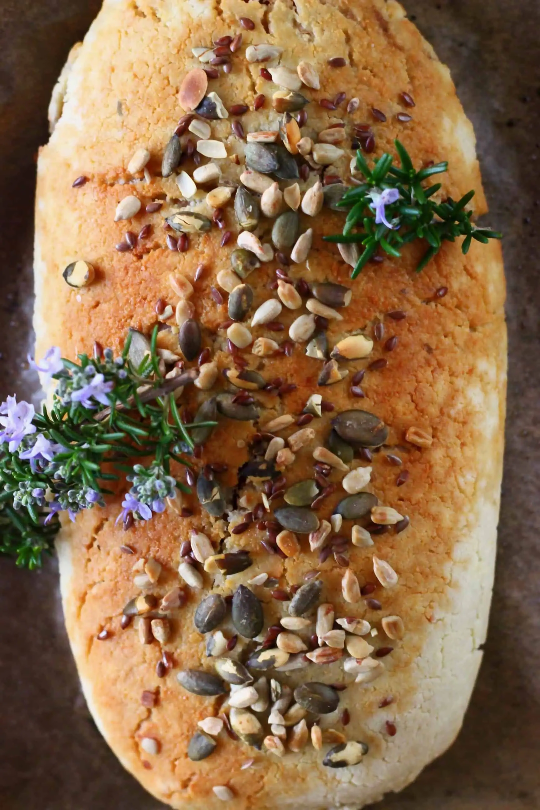 A golden brown vegan wellington topped with mixed seeds and sprigs of rosemary on a baking tray