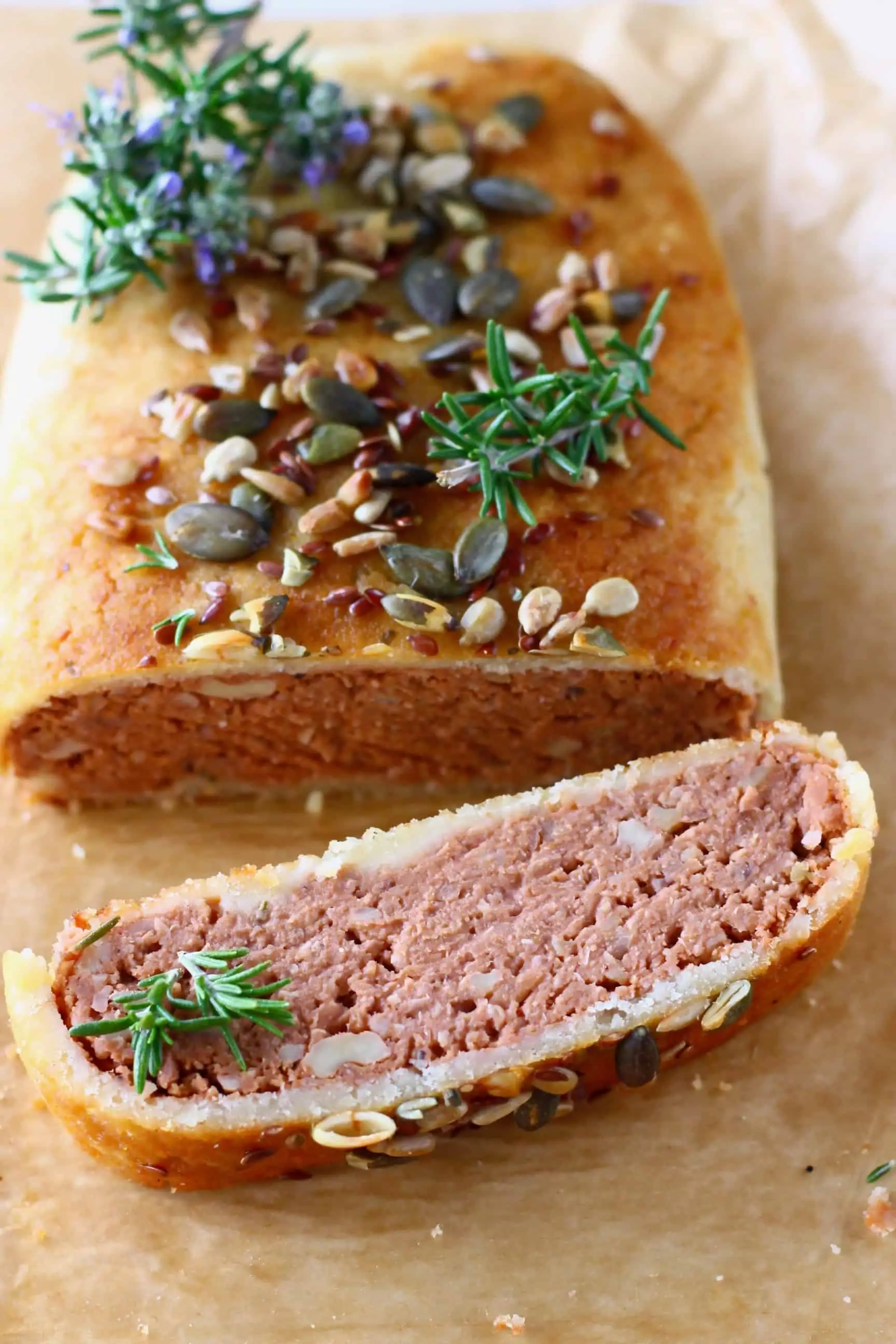 A vegan Wellington topped with mixed seeds with a slice on the side on a sheet of baking paper