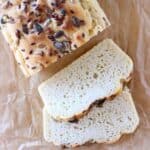 A loaf of gluten-free vegan almond bread sprinkled with mixed seeds with two slices of white bread next to it against a sheet of brown baking paper