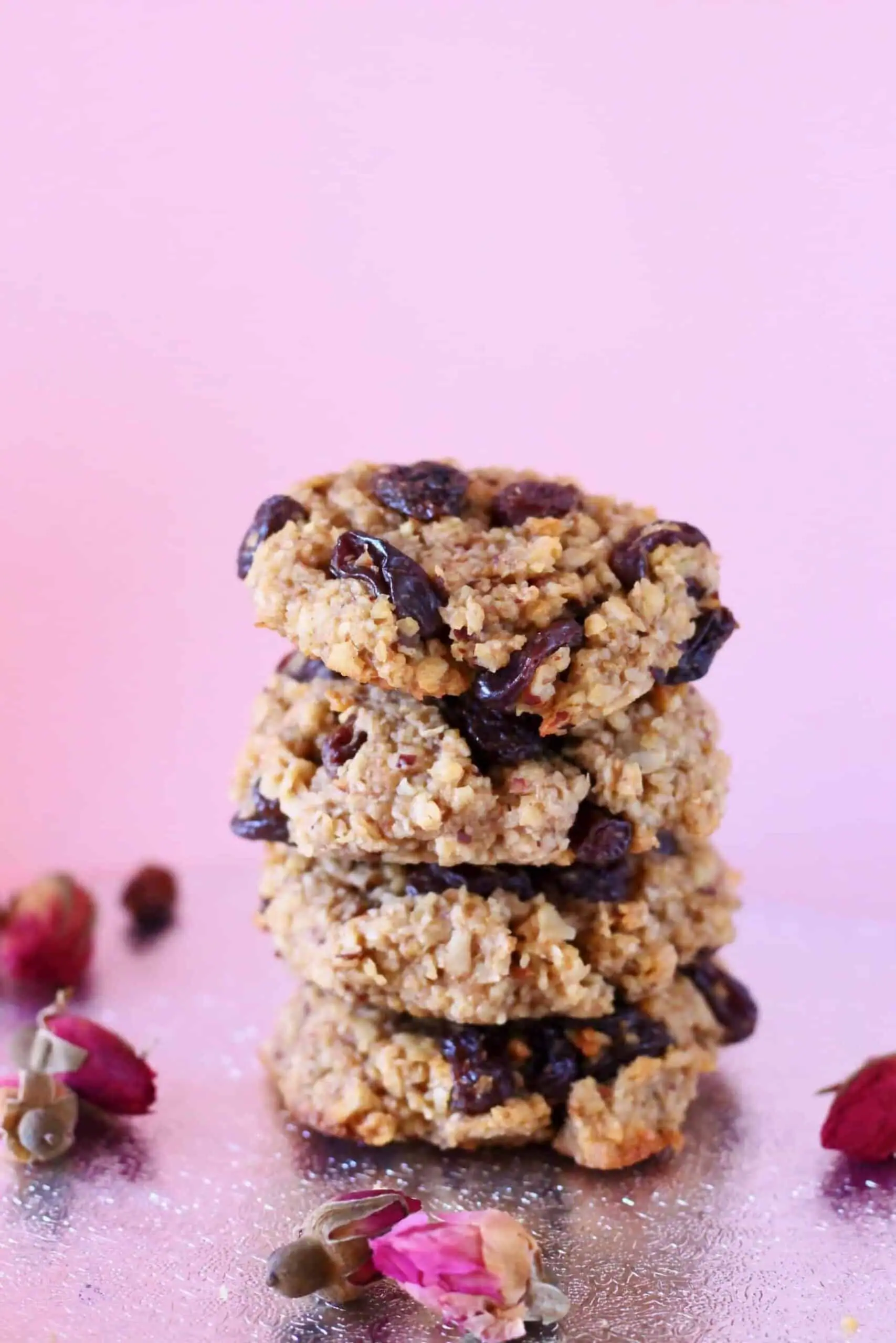 Four oatmeal cookies with raisins stacked up on a silver surface against a light pink background