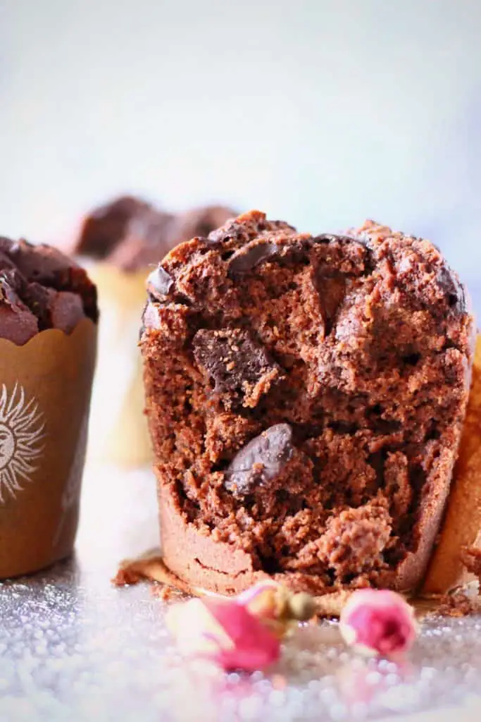 A chocolate muffin with chocolate chips and two other chocolate muffins in the background on a silver surface against a grey background