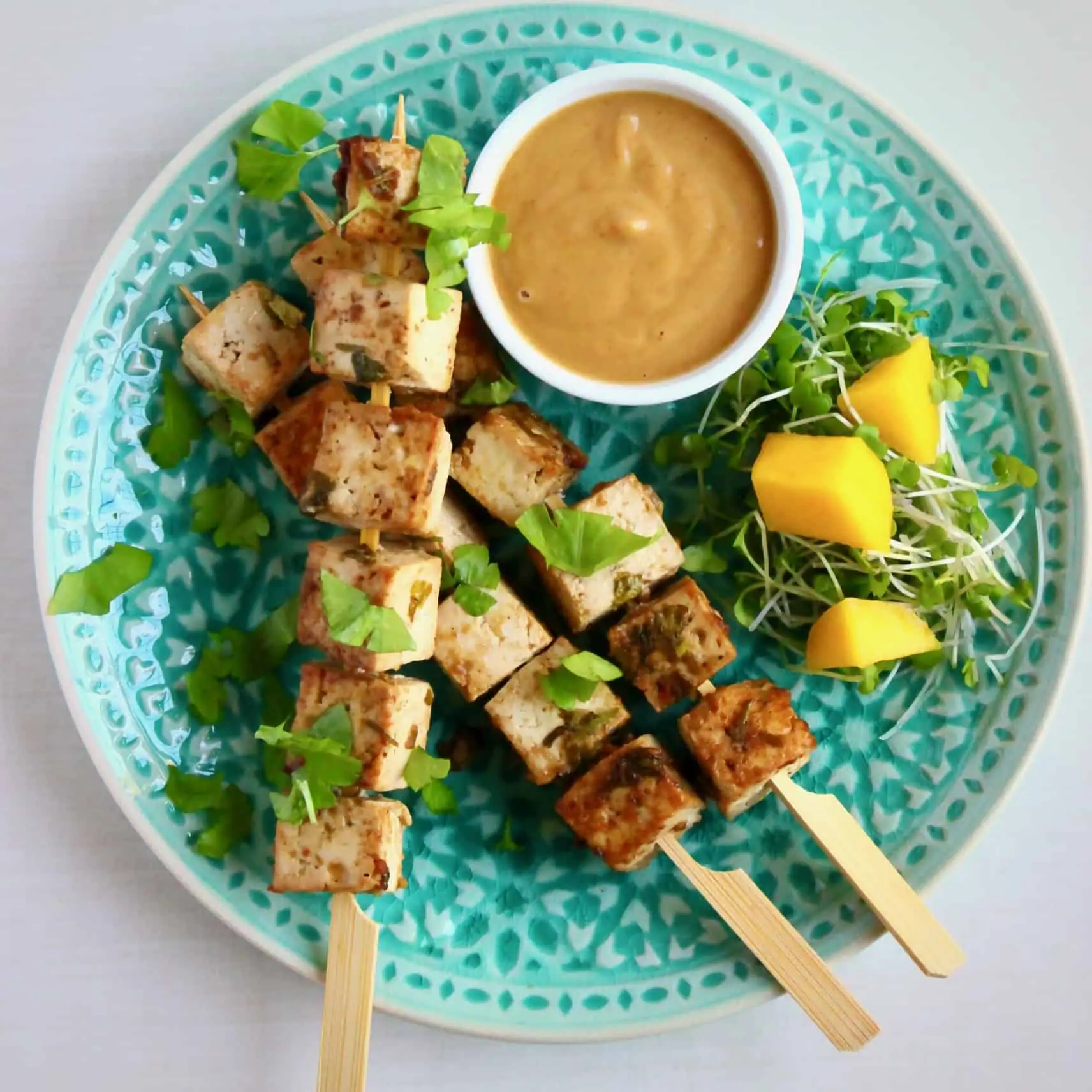 Three skewers with brown tofu cubes on a green plate with a light brown sauce in a white bowl