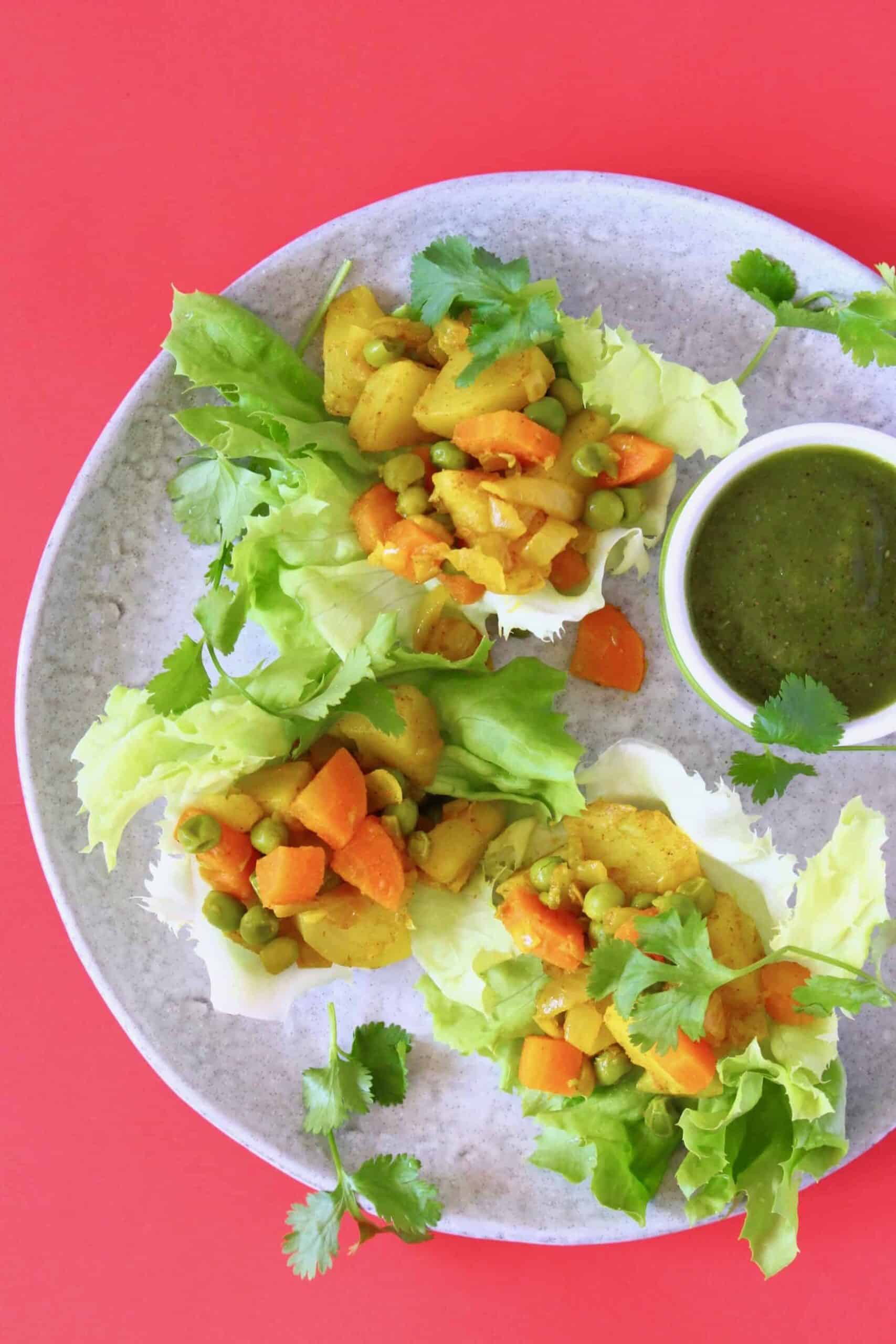 Three lettuce cups filled with curried potato, carrots and green peas on a grey plate with a pot of green chutney against a bright pink background