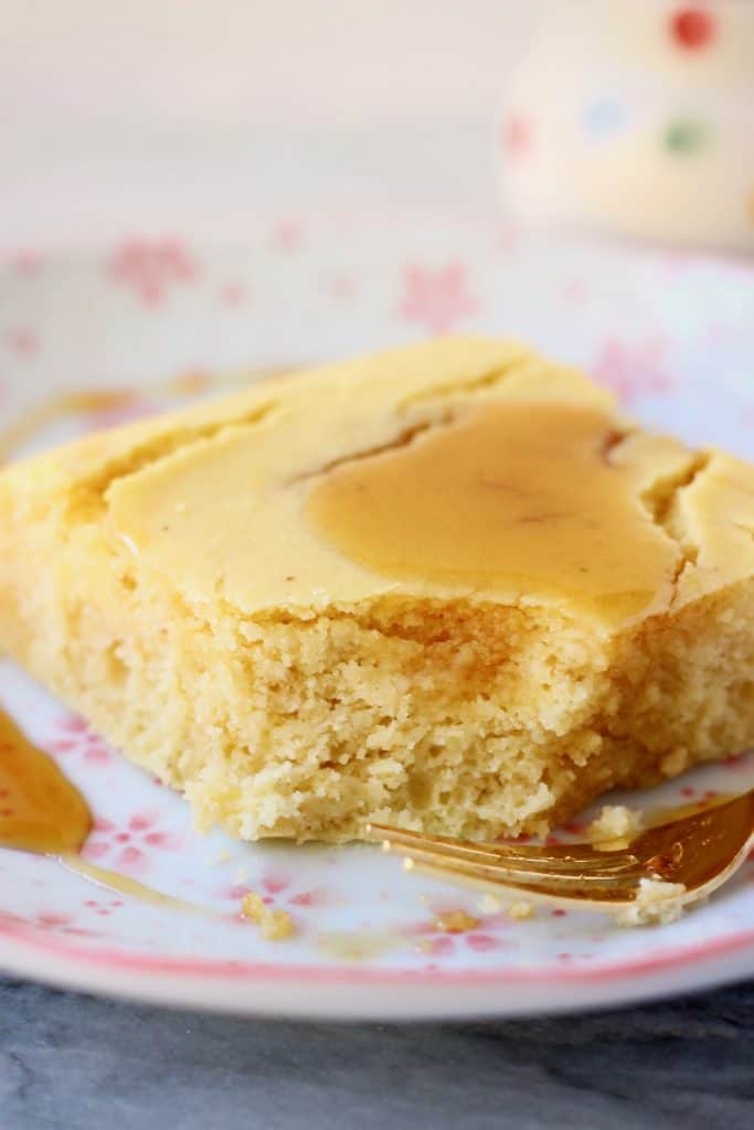 Photo of a square of cornbread with a bite taken out of it drizzled with maple syrup on a white plate with pink flowers and a gold fork