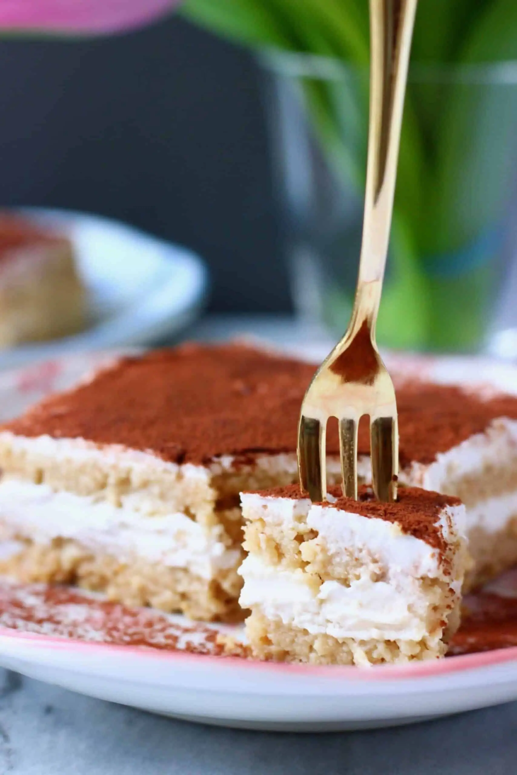 Photo of a slice of tiramisu with a bite taken out of it on a white plate with pink flowers and a gold fork taking a mouthful