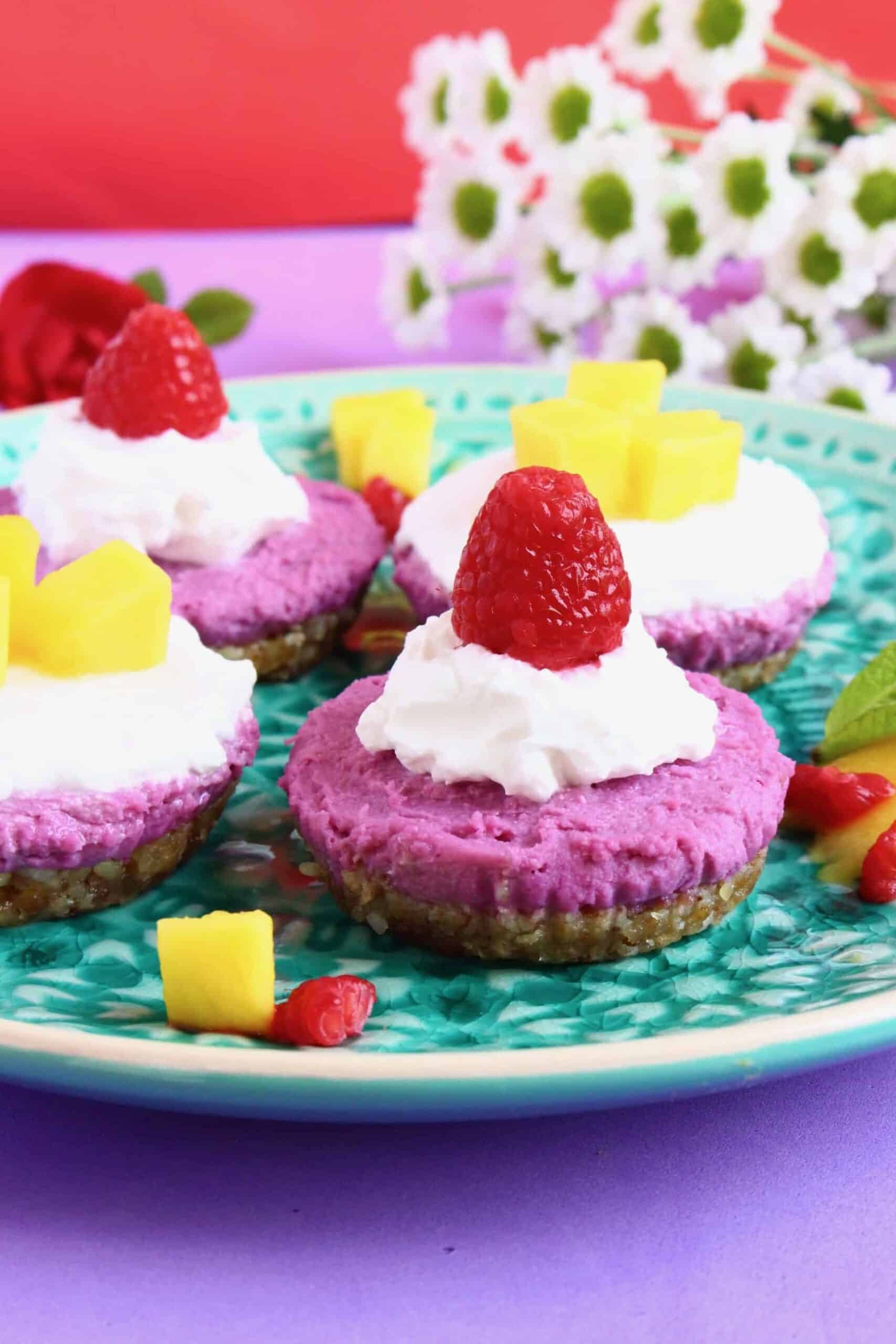 Photo of four purple cheesecakes topped with whipped cream and fruit on a green plate abasing a purple background