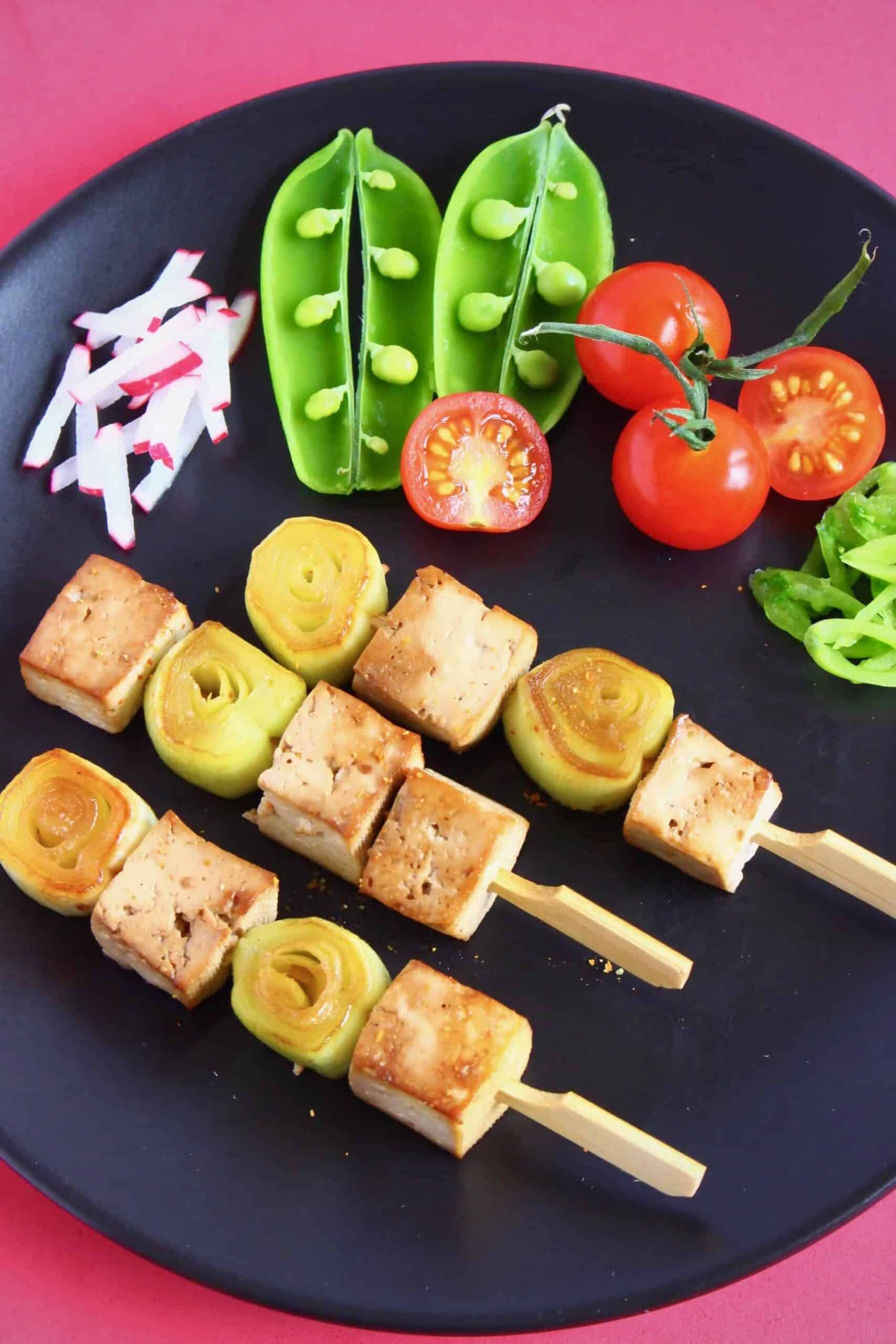 Tofu cubes and sliced leeks on three wooden skewers on a black plate