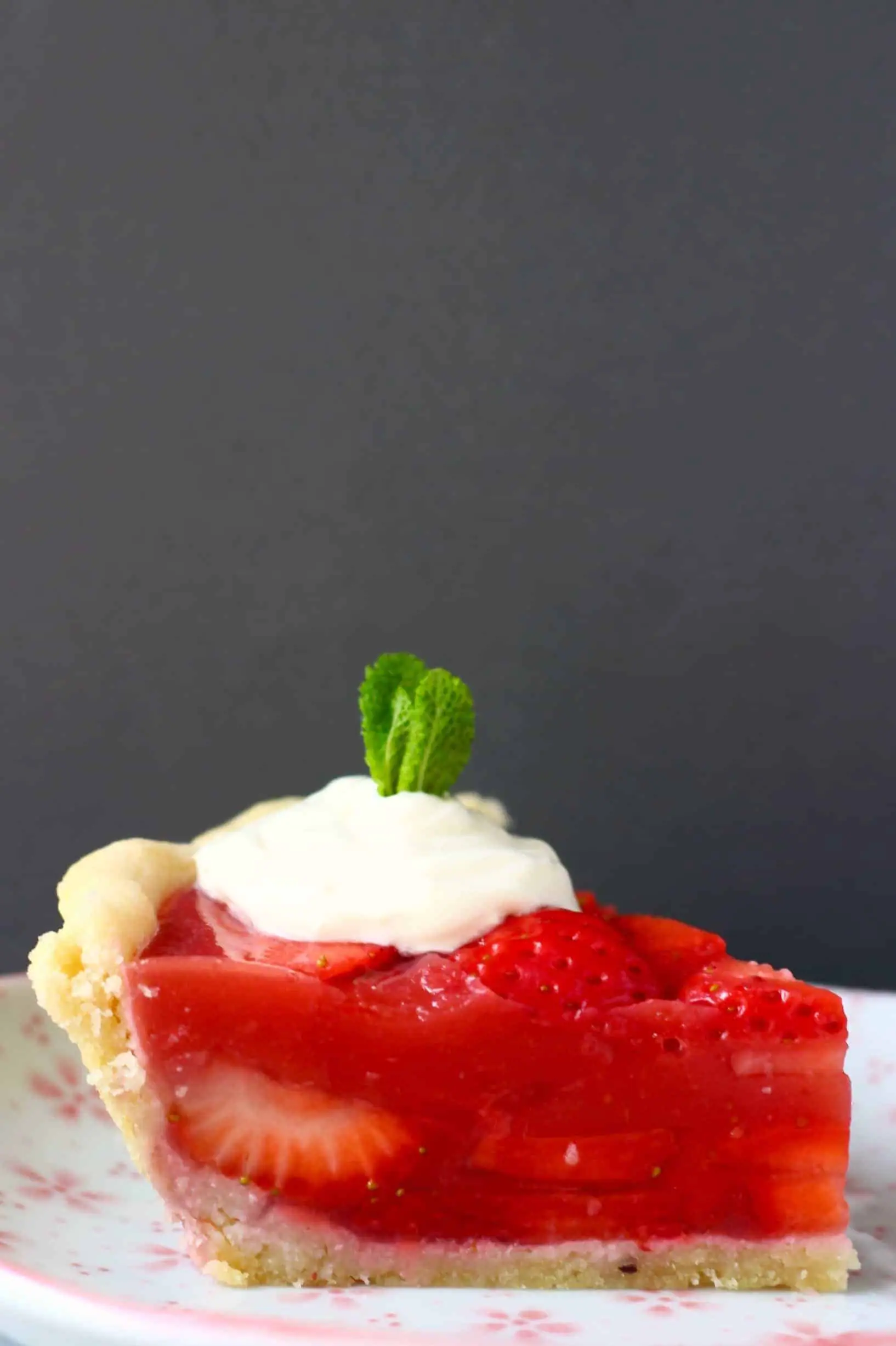 A slice of strawberry pie topped with white cream and a sprig of mint against a grey background
