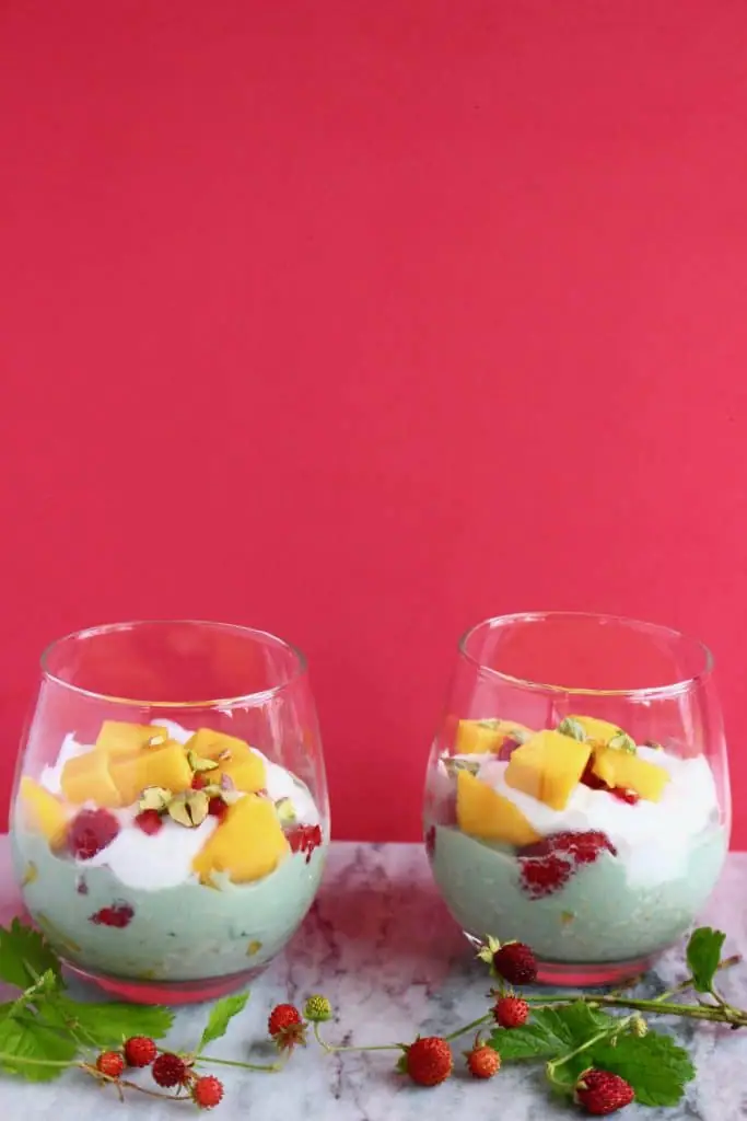 Photo of two glasses filled with green oats and topped with chopped orange fruit and white yogurt against a bright pink background