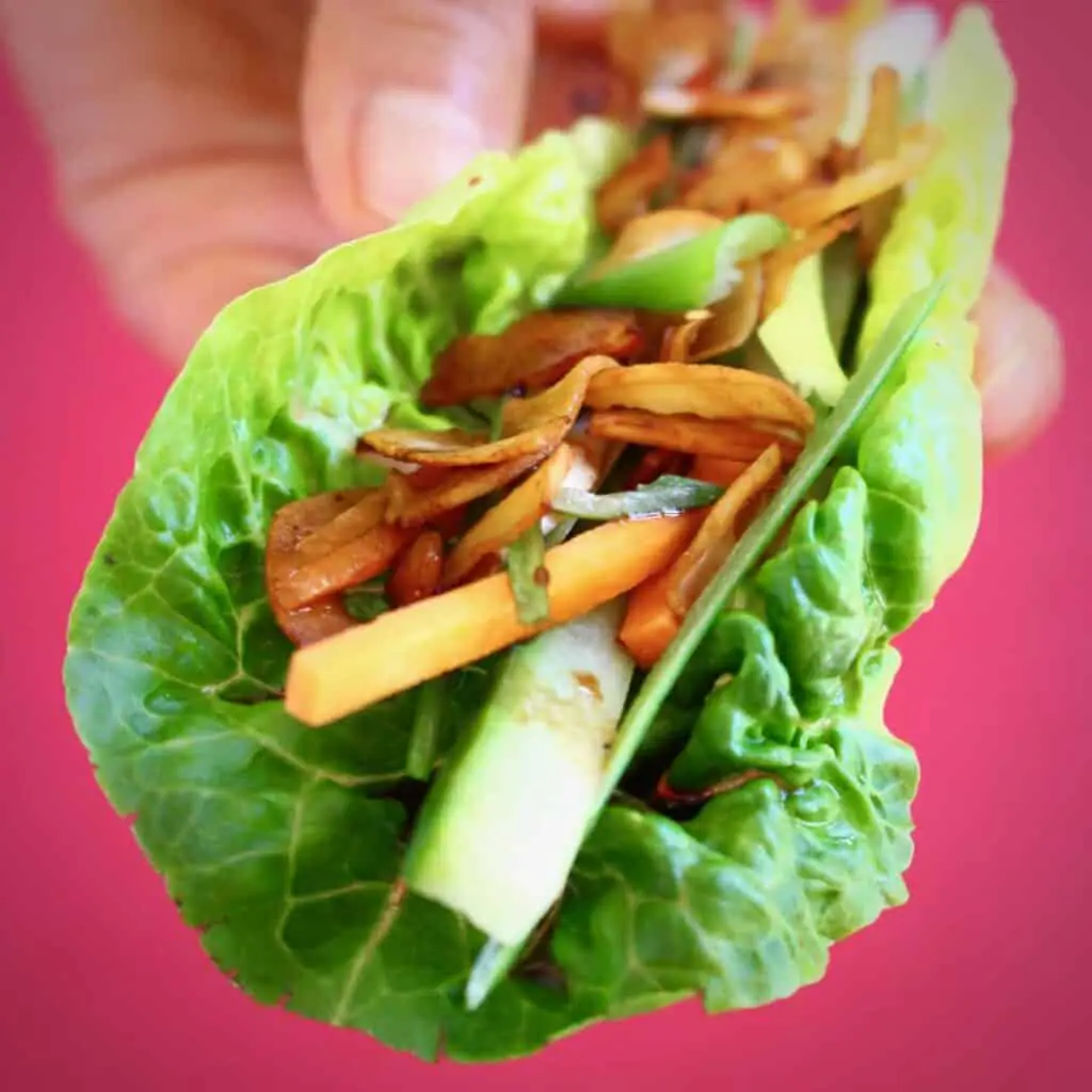 A lettuce wrap filled with brown coconut pieces, sliced spring onions and cucumber held up with a hand against a bright pink background