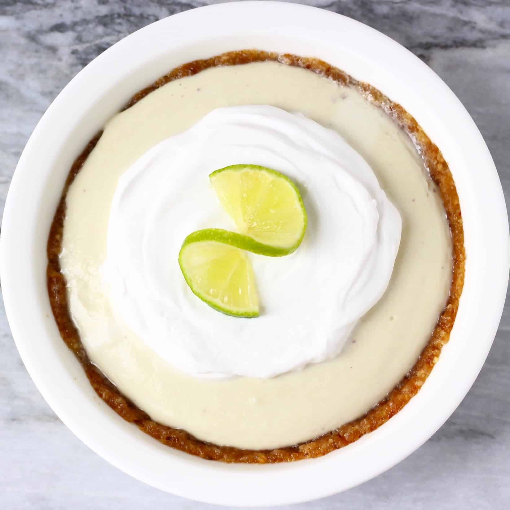 A brown pie crust filled with light green cream topped with white cream and a slice of lime in a white pie dish against a marble background