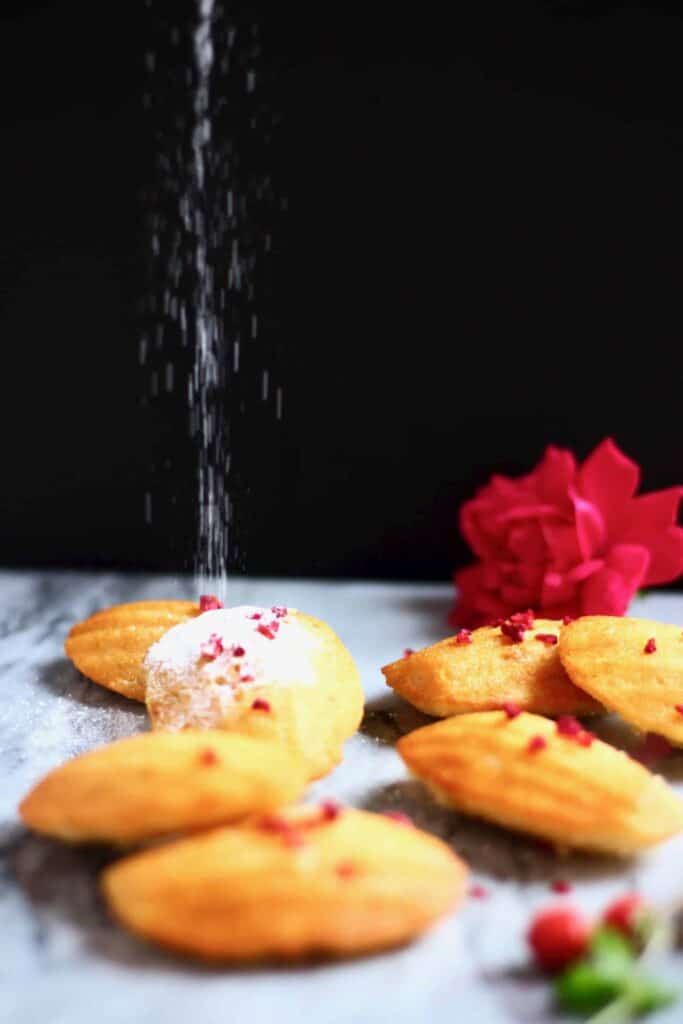 Seven madeleines on a marble surface against a black background with icing sugar being sprinkled over