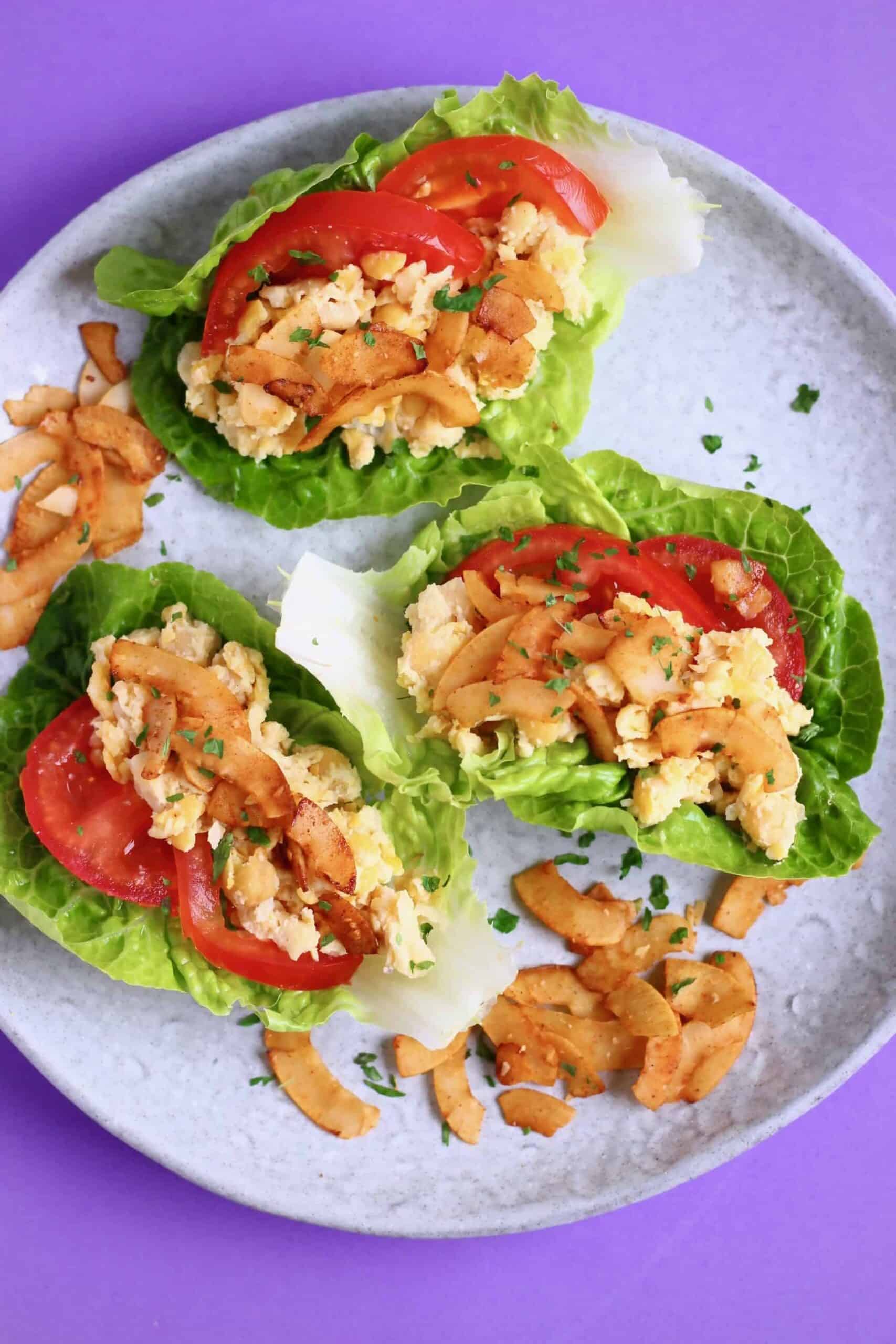 Three lettuce cups filled with creamy chickpeas, coconut bacon and tomatoes on a grey plate against a purple background
