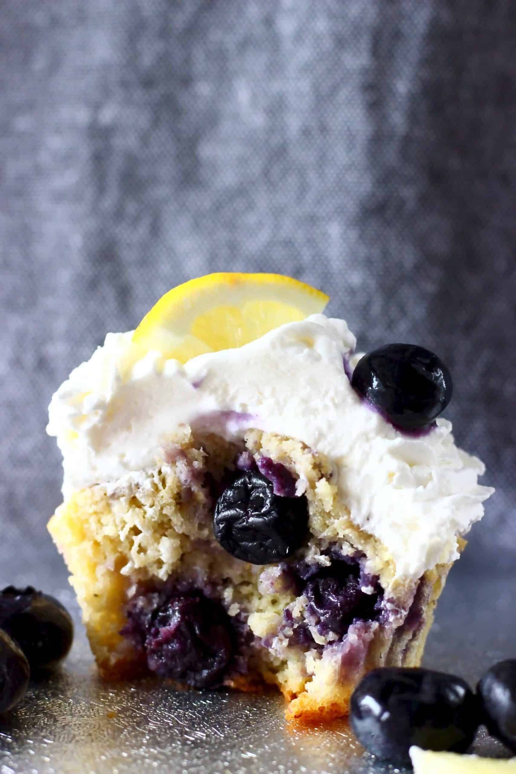 A blueberry cupcake with a bite taken out of it topped with creamy frosting and a lemon wedge against a grey background
