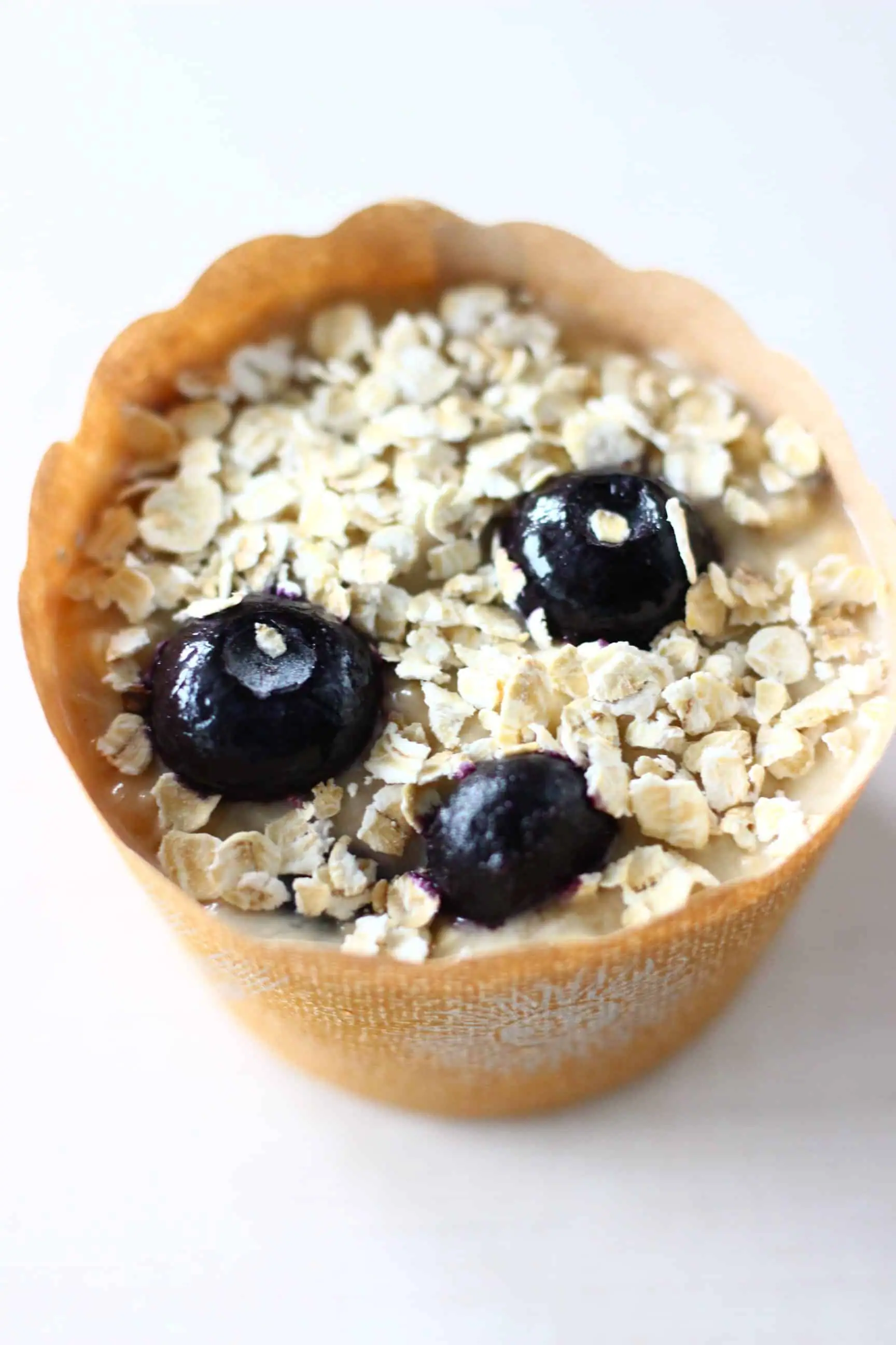 A brown muffin case filled with raw gluten-free vegan blueberry oatmeal muffin batter sprinkled with oats
