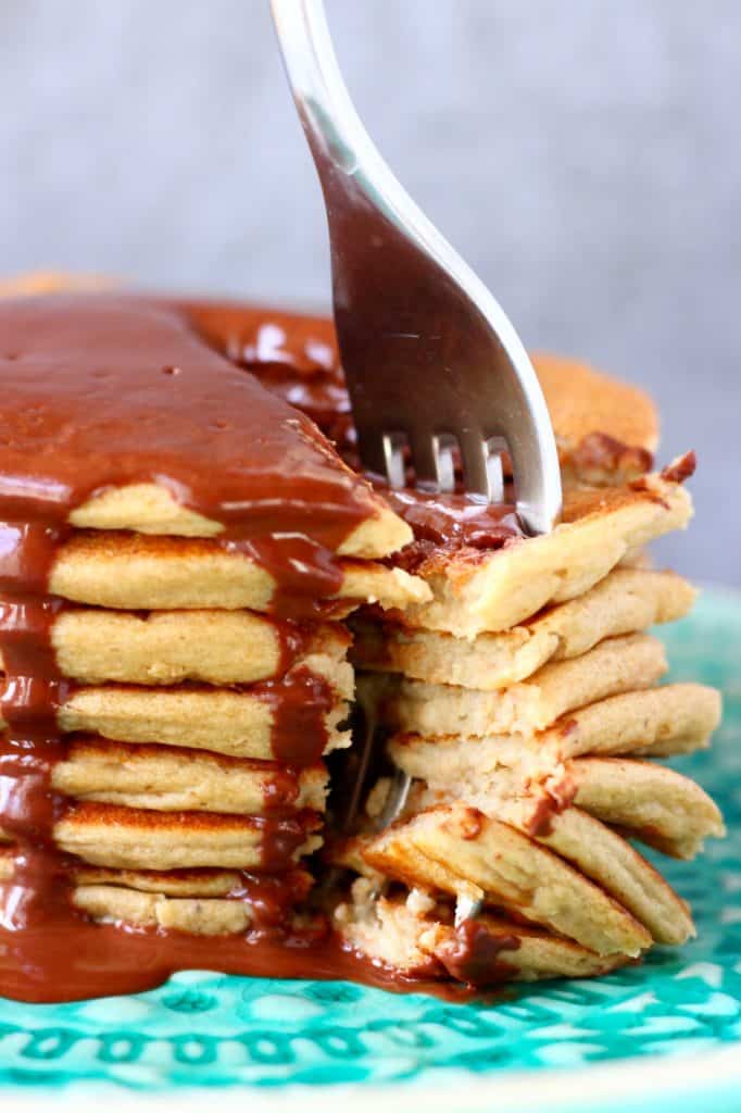 Photo of a stack of pancakes covered in chocolate sauce with a portion cut out of it and a silver fork through it on a green plate against a grey background