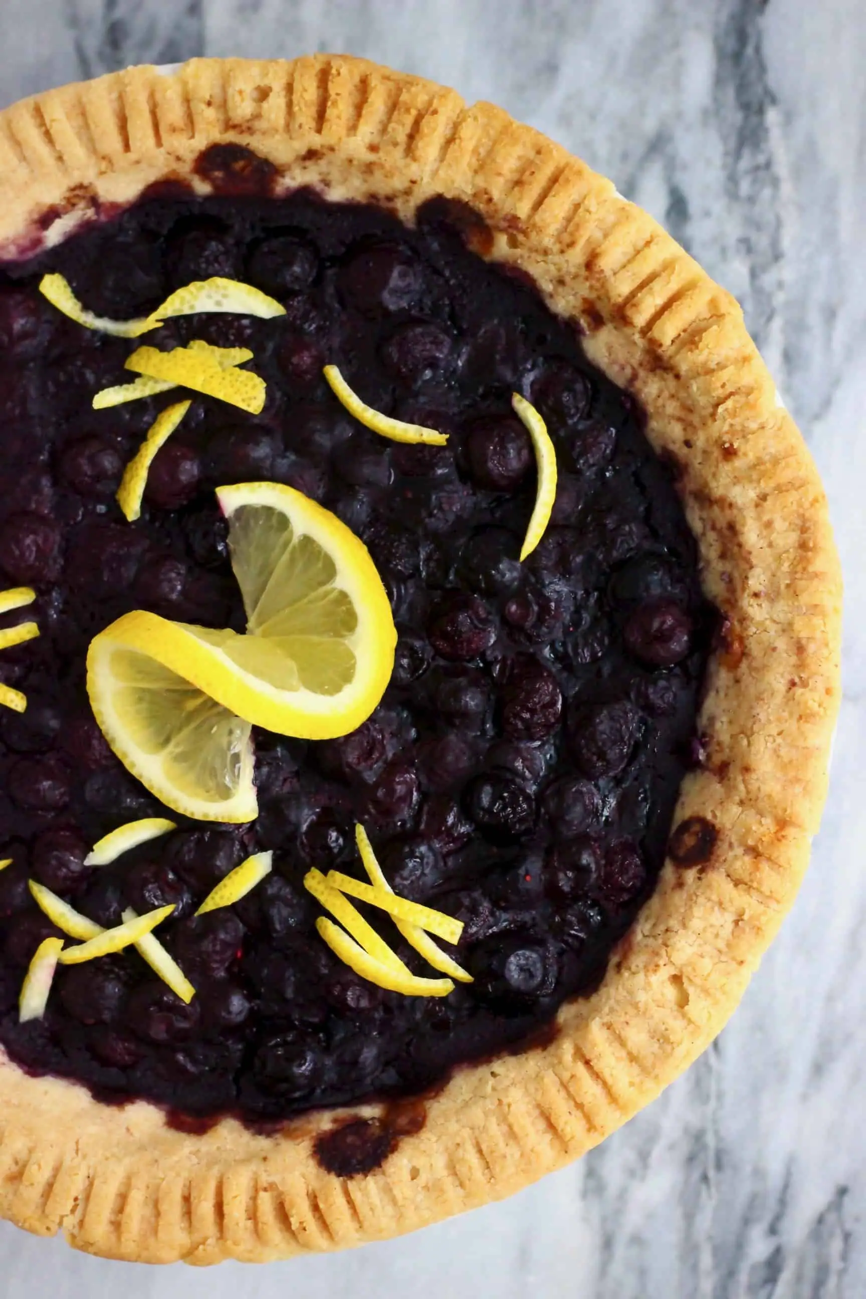 A vegan blueberry pie decorated with lemon peel and a slice of lemon against a marble background