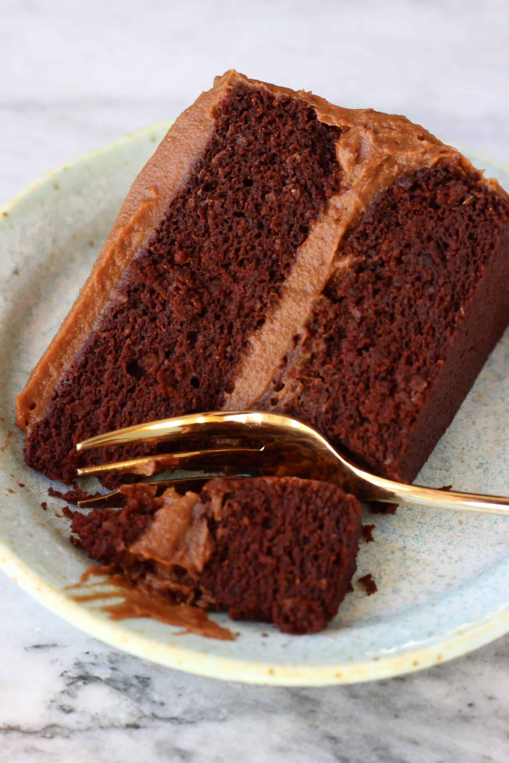 A slice of gluten-free vegan chocolate cake with chocolate frosting on a blue plate with a gold fork 