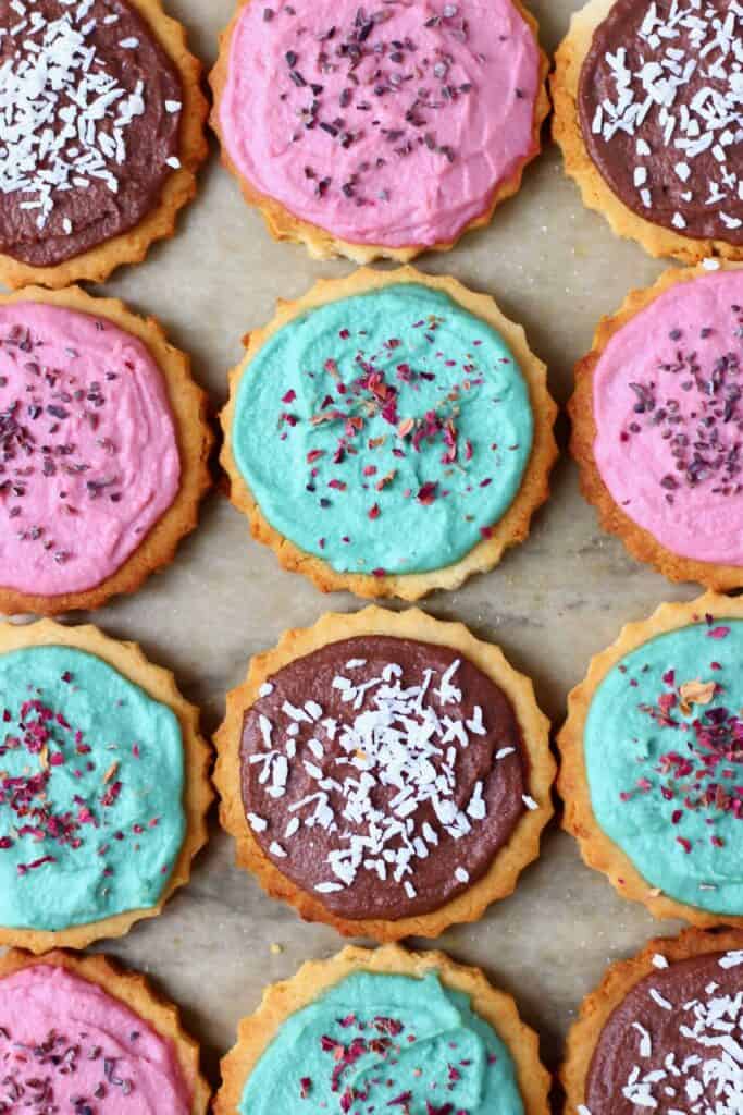 Photo of twelve circular cookies topped with different coloured frosting and sprinkles on a sheet of brown baking paper