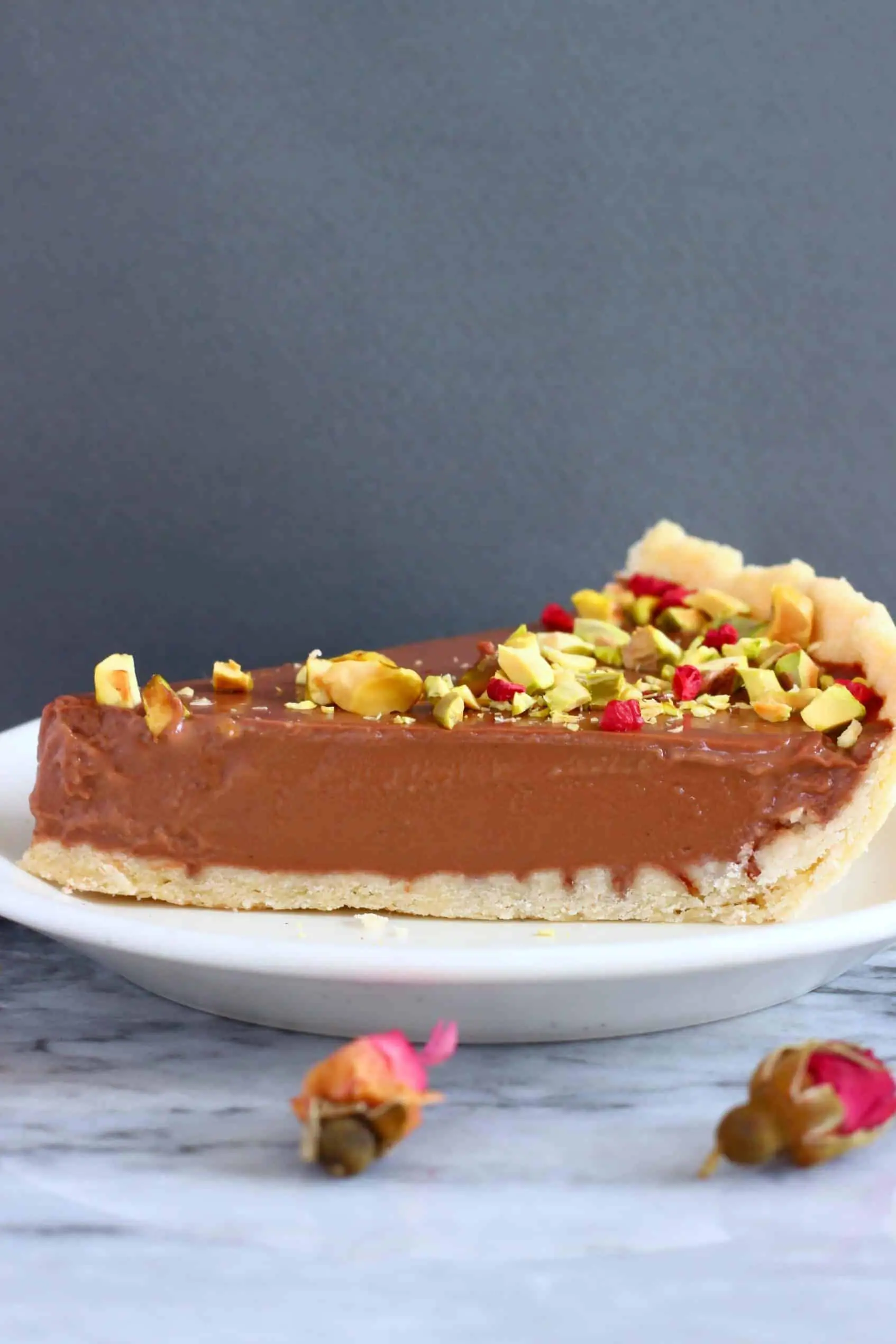 A slice of chocolate tart topped with chopped pistachios and freeze-dried raspberries on a white plate against a grey background